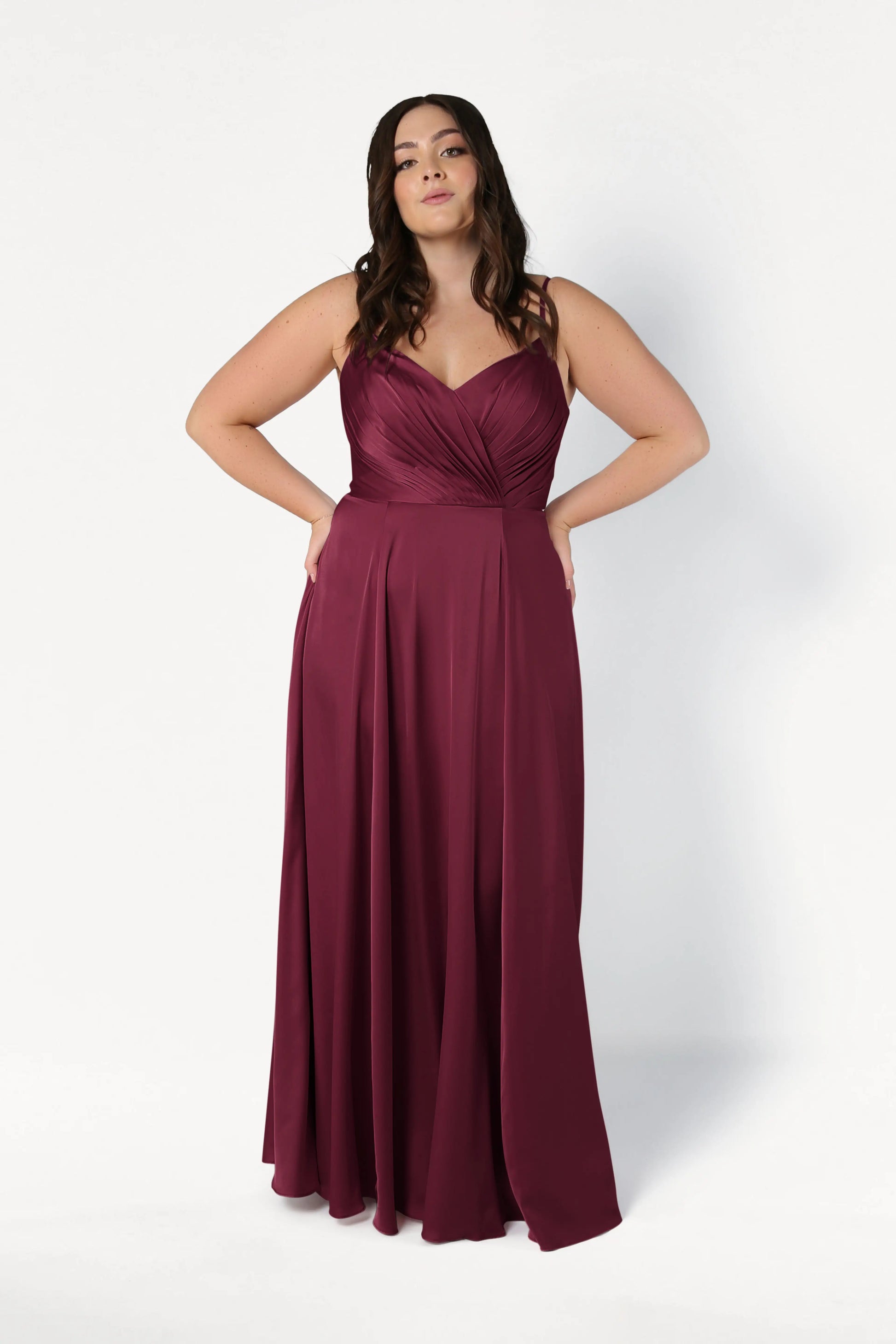 A plus-size model wears the TH&TH Isla Bridesmaid Dress in Merlot Satin, featuring a pleated sweetheart bodice, spaghetti straps, and a flowing floor-length A-line skirt, posing against a white background.
