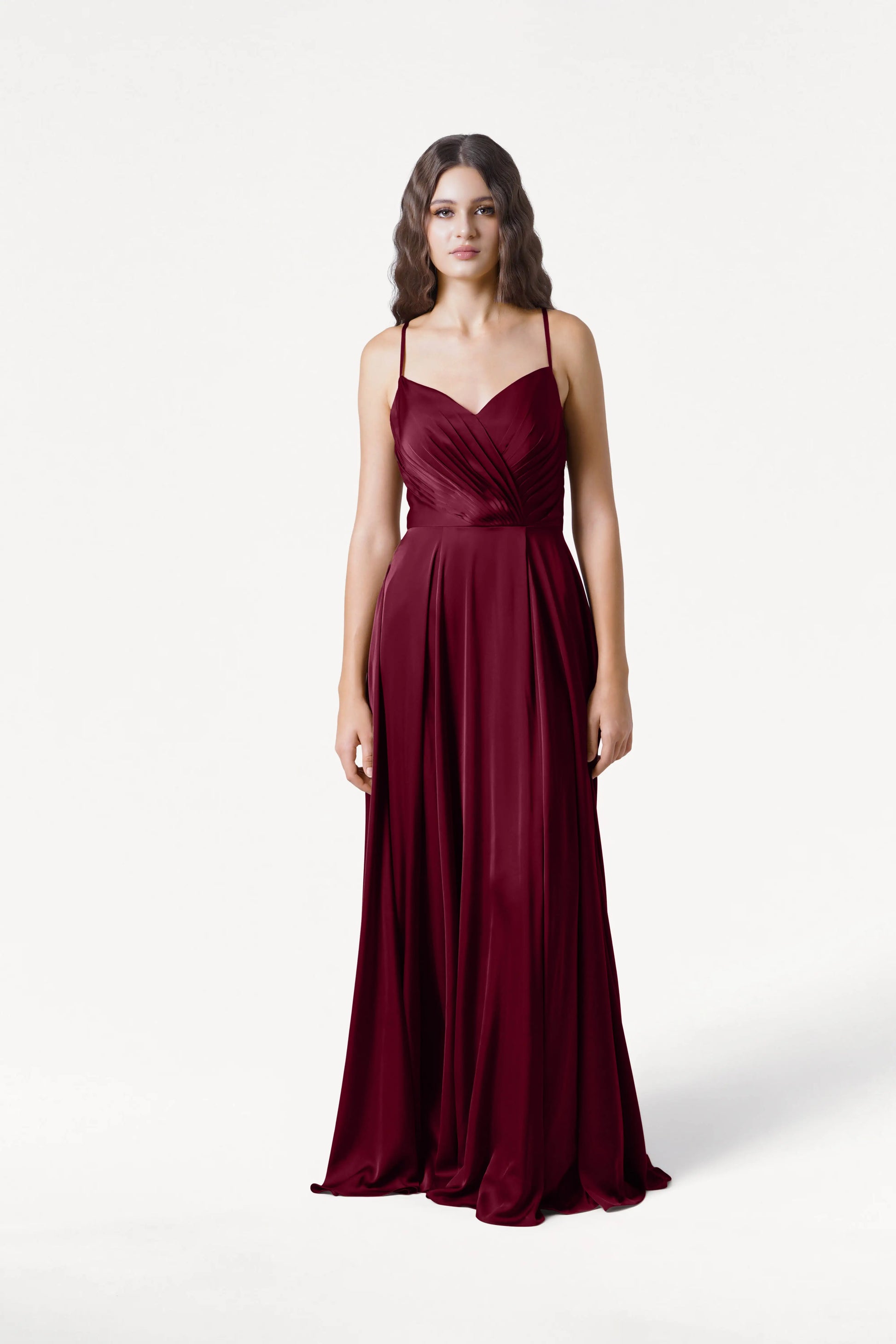 A woman faces forward in the floor-length Isla Bridesmaid Dress by TH&TH, crafted in merlot satin with spaghetti straps and a pleated bodice, set against a plain white background.