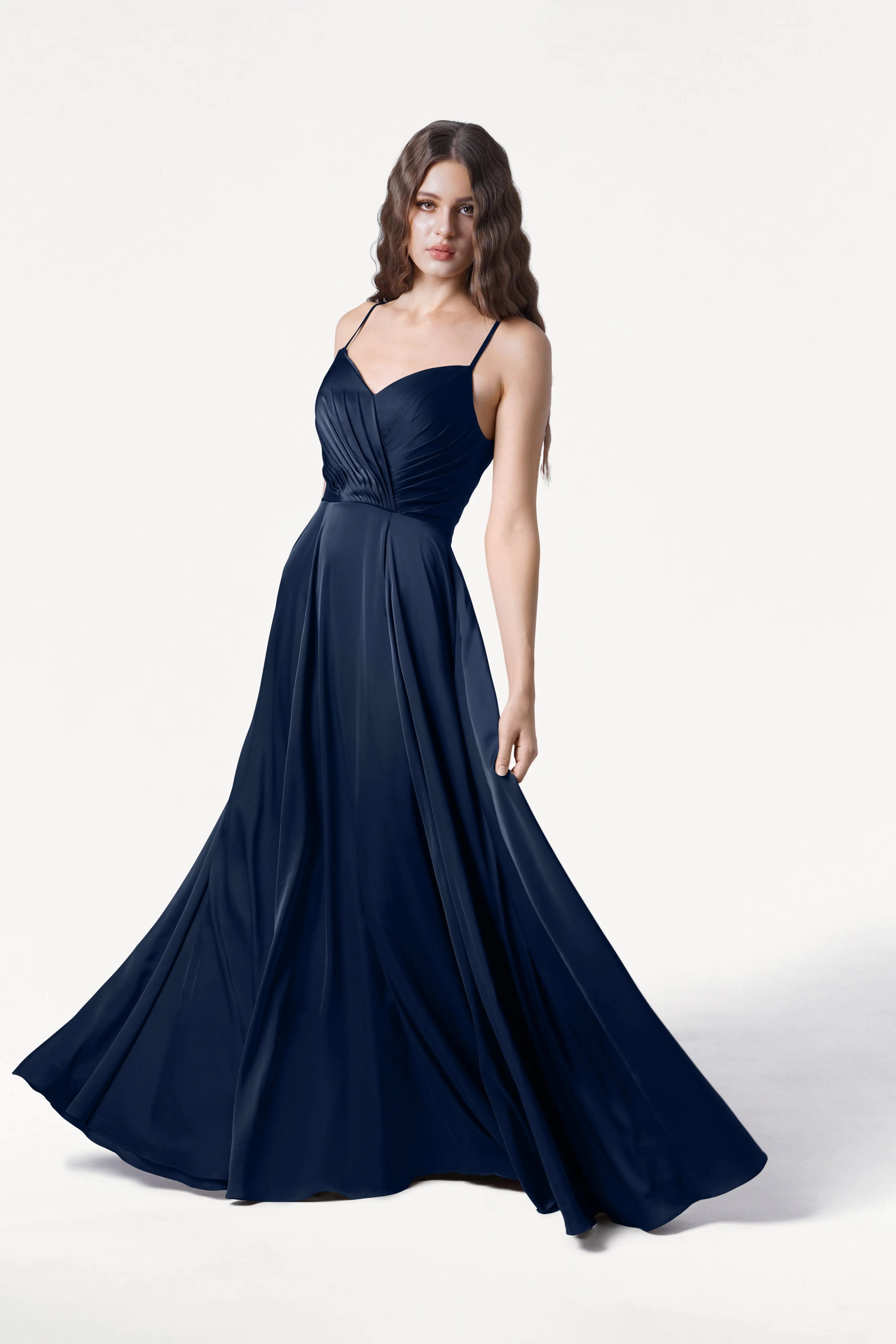 A woman wears the Isla Bridesmaid Dress in Navy Satin by TH&TH—a sleeveless, floor-length gown with a fitted pleated bodice, adjustable straps, and a flared skirt—posing against a white background for an elegant look.