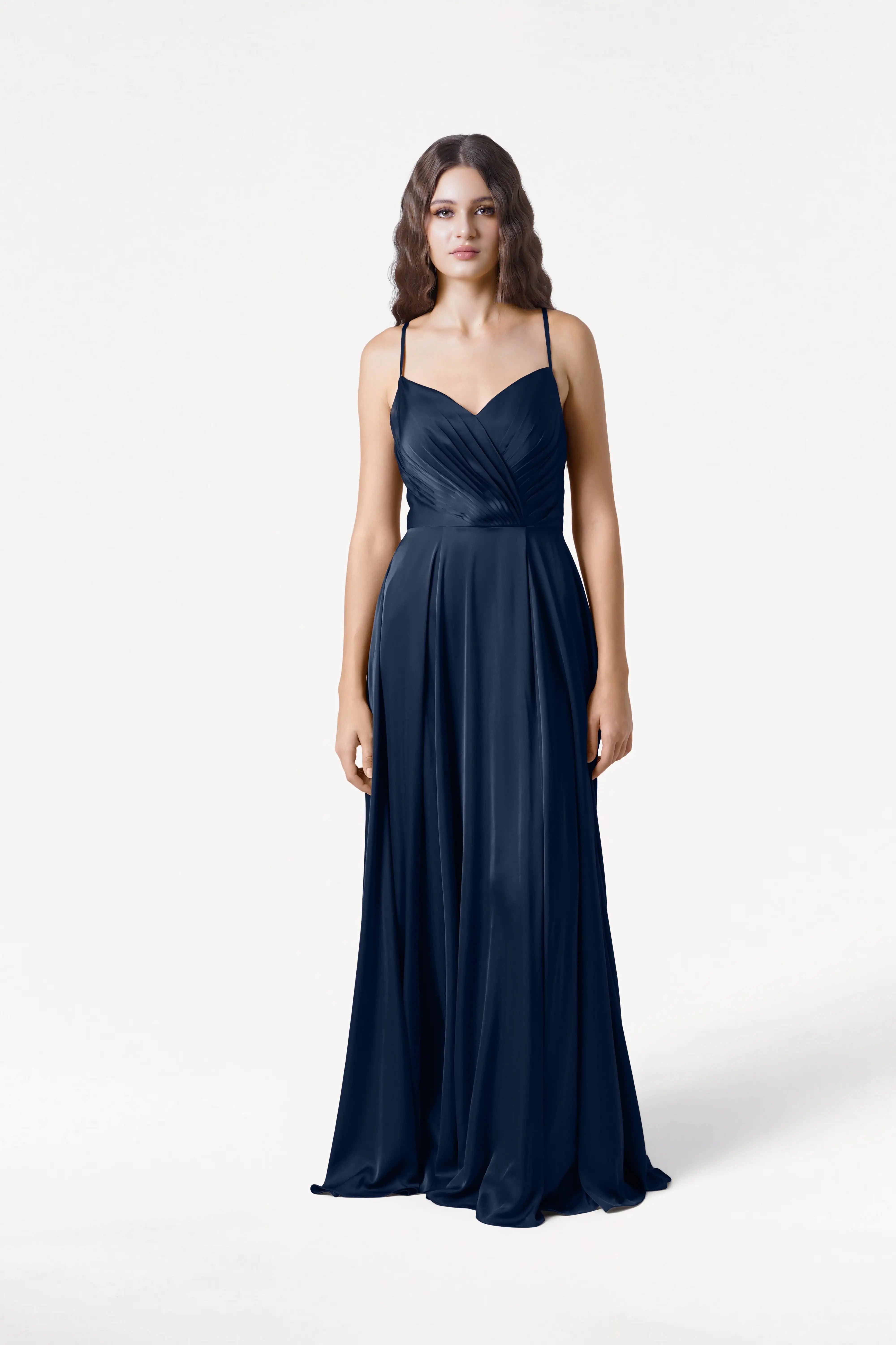 A woman with wavy brown hair wears the Isla Bridesmaid Dress in Navy Satin by TH&TH, featuring adjustable straps and a gathered bodice, standing against a plain white background.