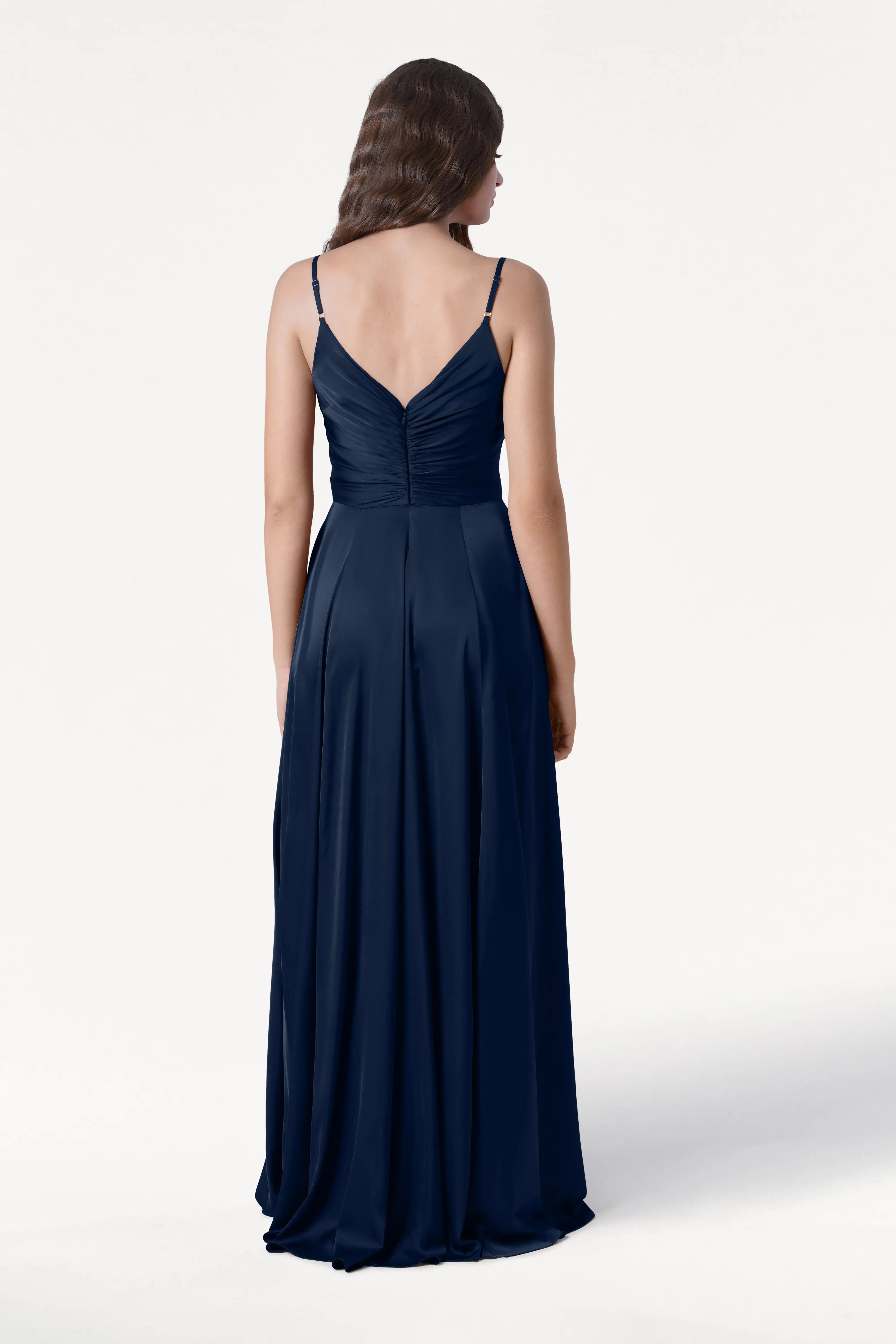 A woman with long, wavy hair stands with her back to the camera in the TH&TH Isla Bridesmaid Dress in Navy Satin, featuring a pleated bodice and adjustable straps. She is set against a plain white background.