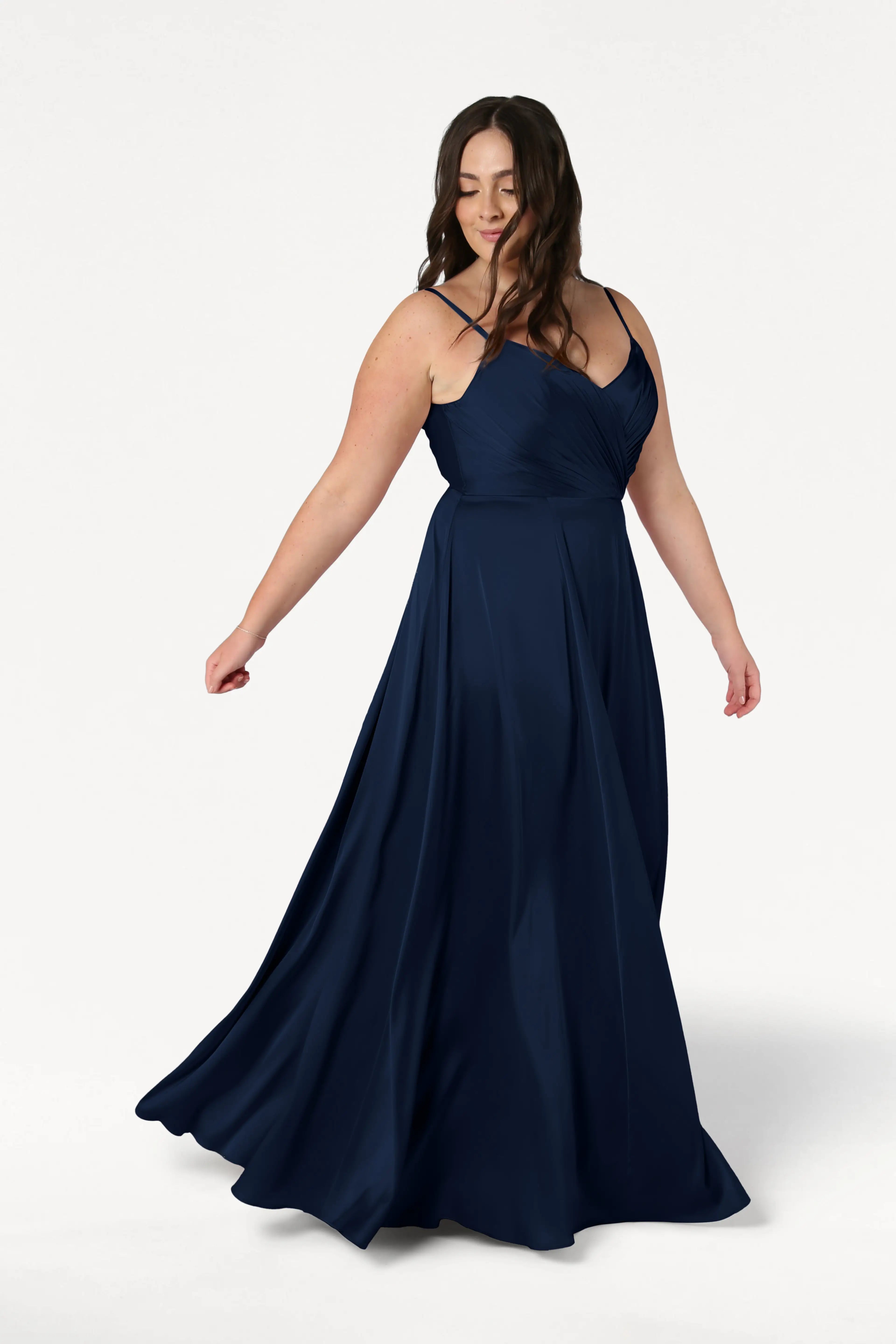 A woman twirls gracefully in the Isla Bridesmaid Dress in Navy Satin by TH&TH, featuring a pleated bodice and thin adjustable straps, her long brown hair falling in loose waves against a plain white background.