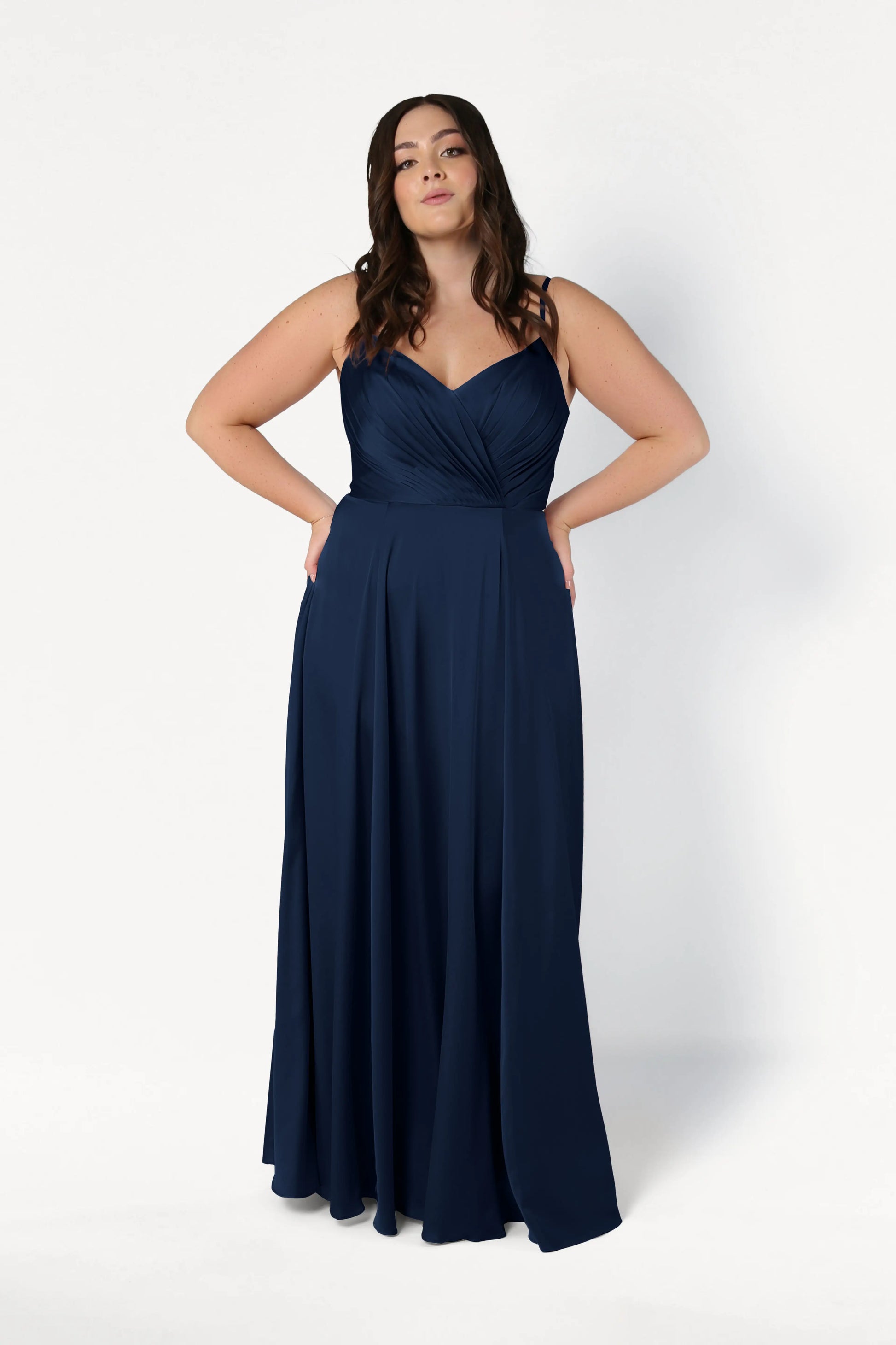 A woman models the Isla Bridesmaid Dress in Navy Satin by TH&TH—featuring a floor-length silhouette, fitted pleated bodice, and adjustable straps—while standing against a plain white background, confidently facing the camera.