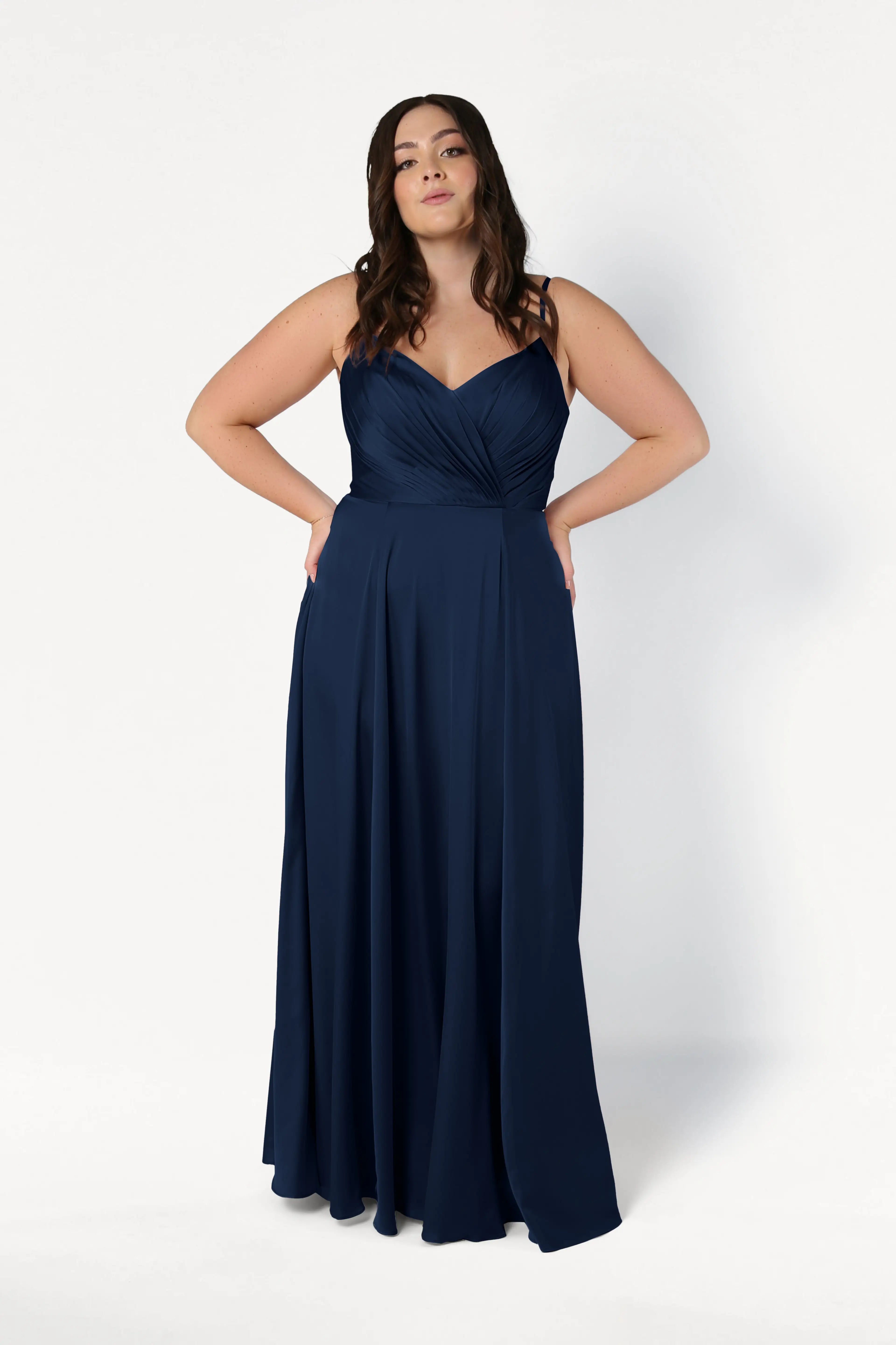 A woman models the Isla Bridesmaid Dress in Navy Satin by TH&TH—featuring a floor-length silhouette, fitted pleated bodice, and adjustable straps—while standing against a plain white background, confidently facing the camera.