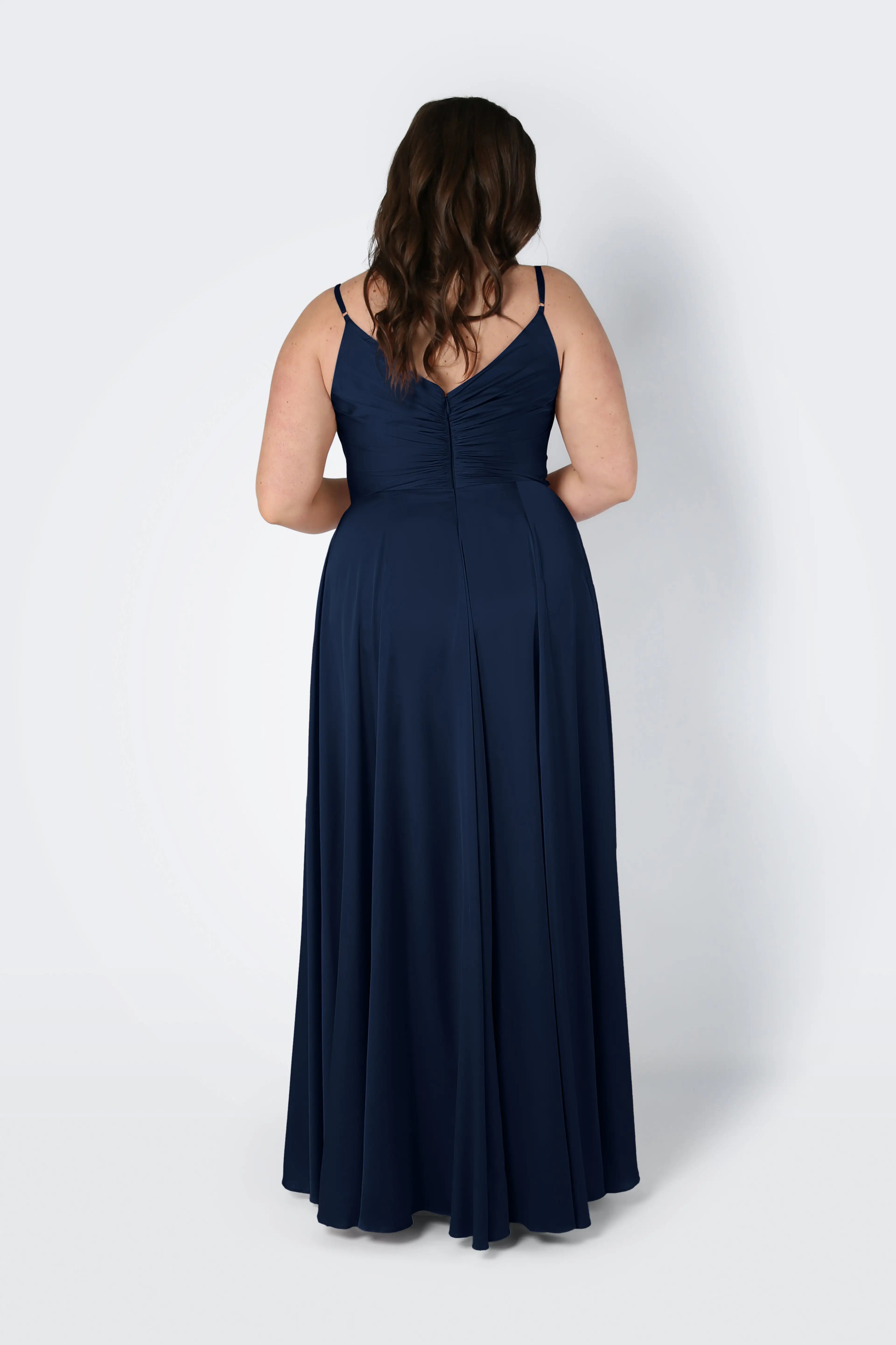 A woman with long brown hair, seen from behind, wears the TH&TH Isla Bridesmaid Dress in Navy Satin. The floor-length dress features adjustable straps, a pleated bodice with gathered detailing, and is set against a plain light gray background.