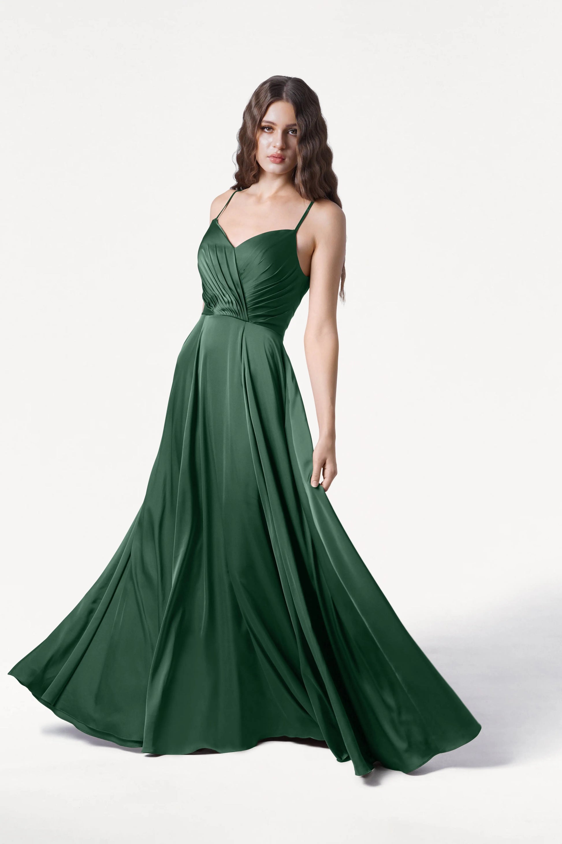 A woman poses against a plain white background in the Isla Bridesmaid Dress in Olive Grove Satin by TH&TH, a floor-length dark green satin gown with thin straps, a fitted pleated bodice, and a gracefully flared skirt.