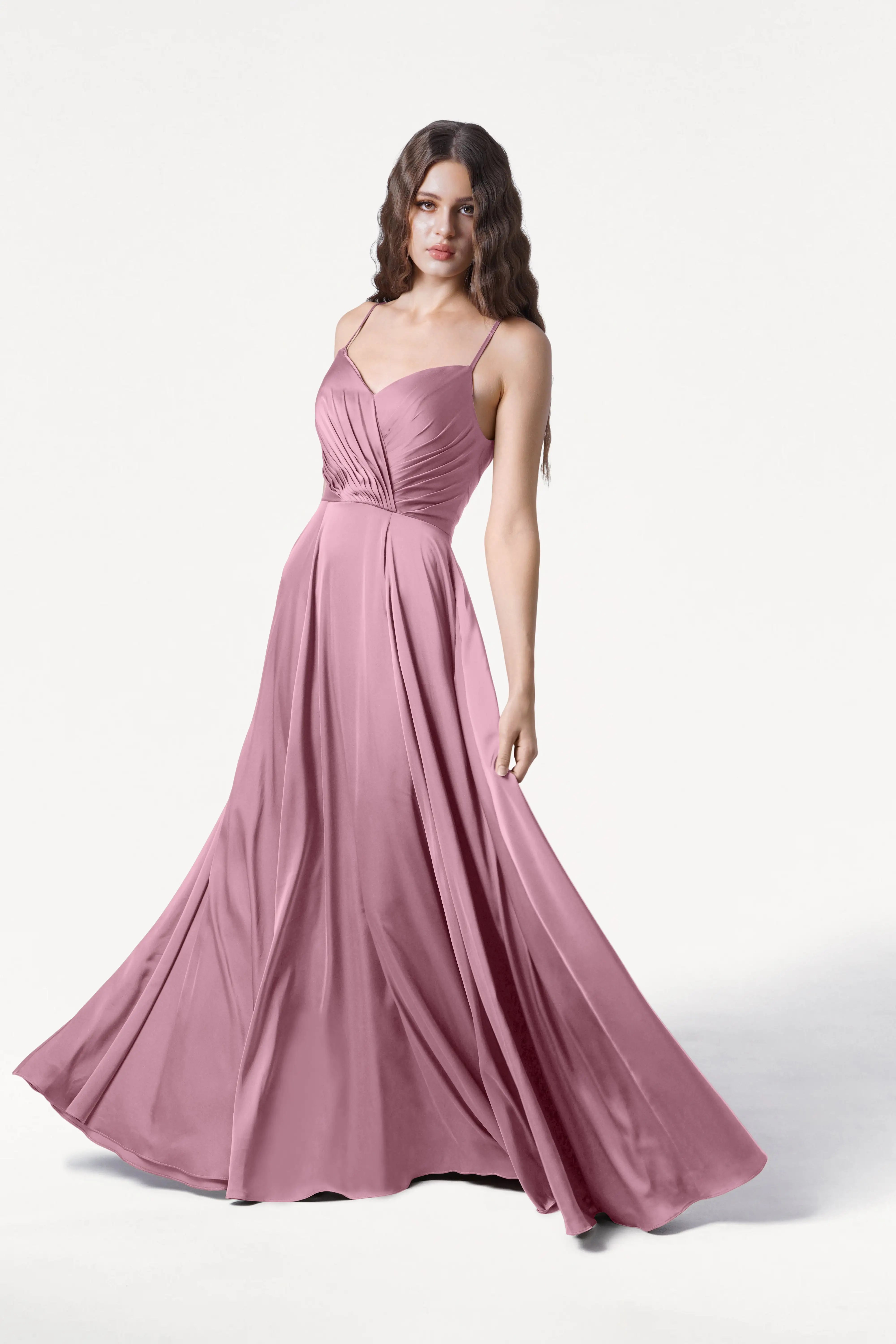 A woman stands against a plain white background wearing the TH&TH Isla Bridesmaid Dress in rose pink satin, featuring a flowing, floor-length silhouette, adjustable straps, and a fitted ruched bodice.