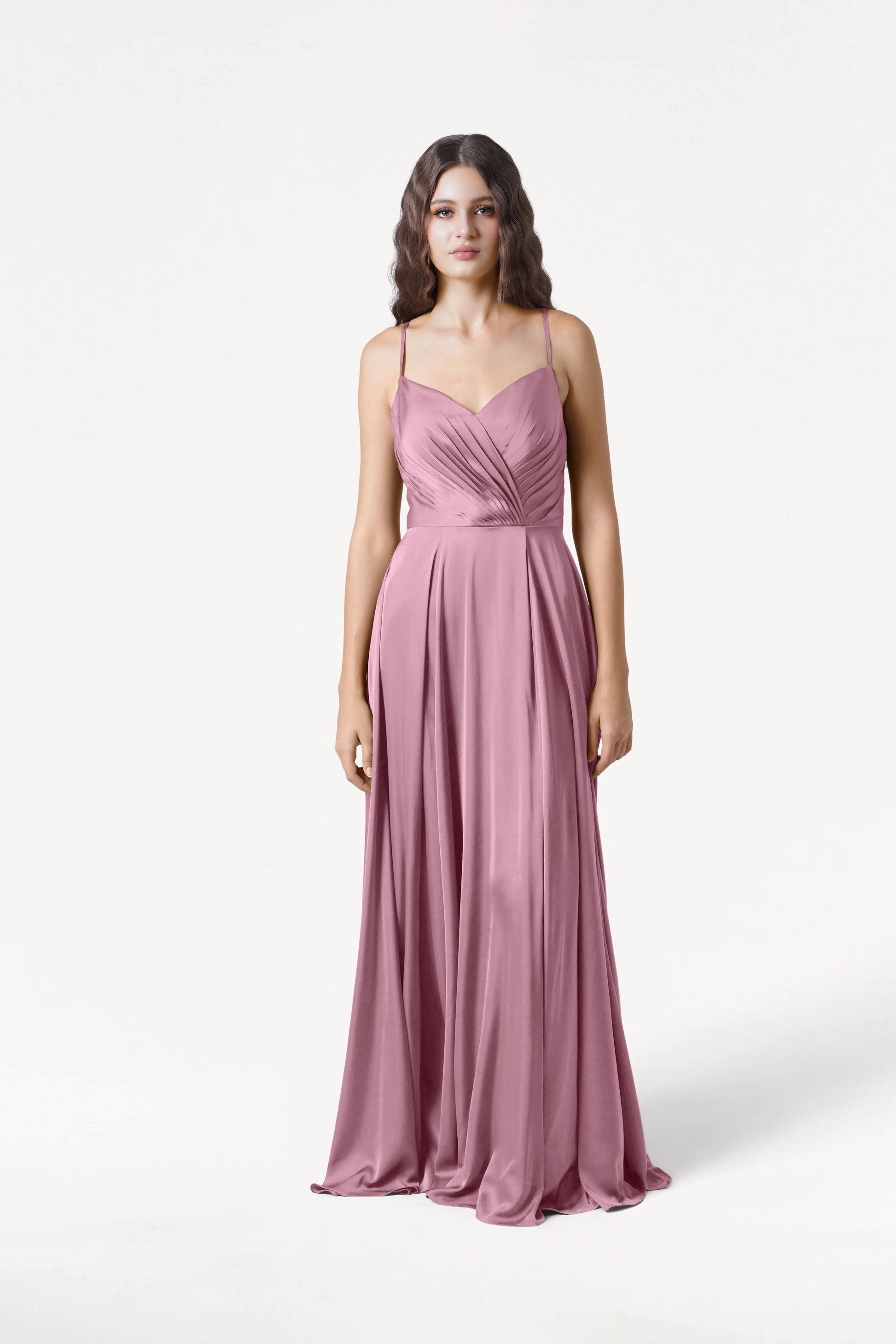 A woman faces forward in the Isla Bridesmaid Dress by TH&TH, a sleeveless, floor-length rose pink satin gown with adjustable spaghetti straps and a pleated bodice, set against a plain white background.