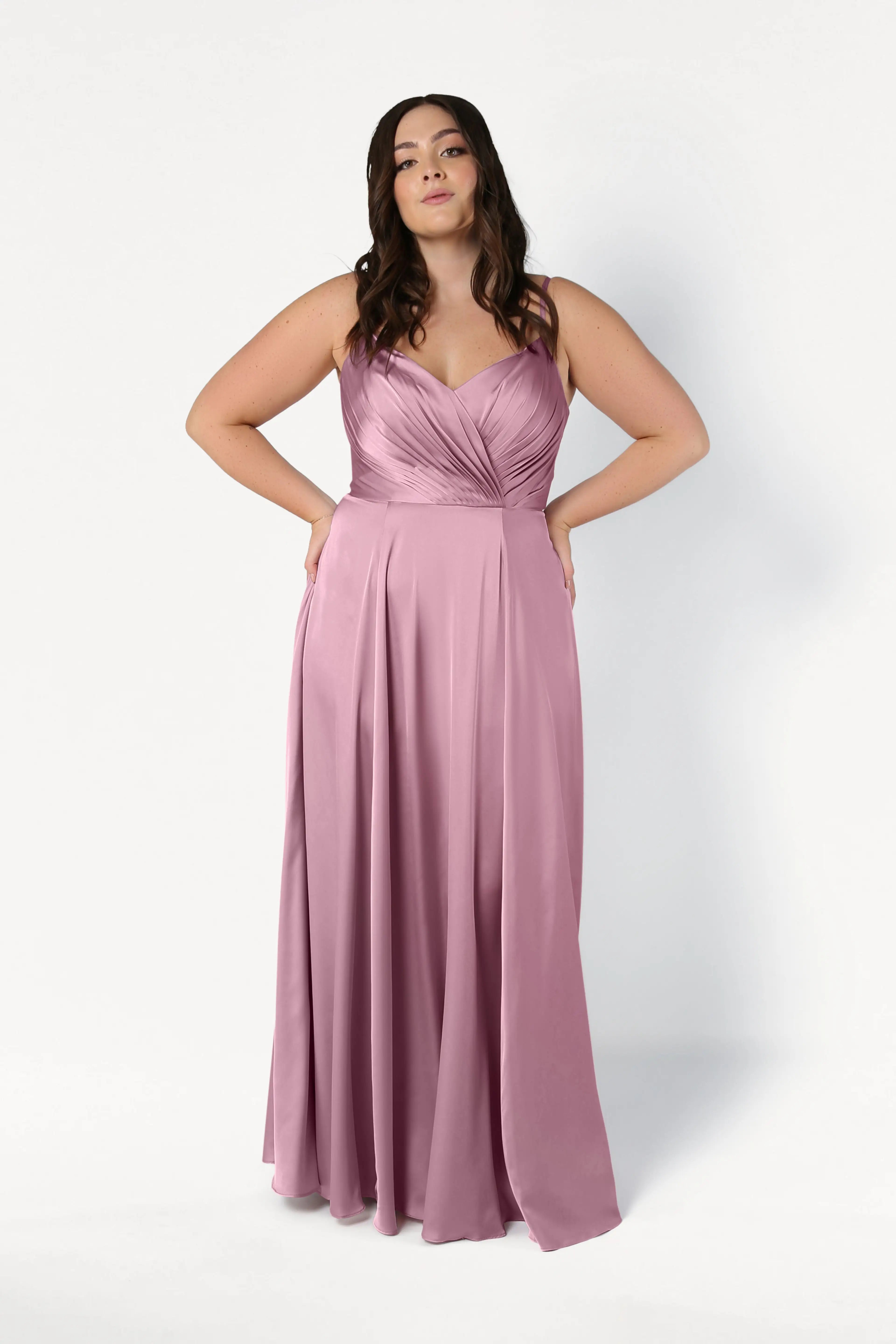 A woman models the TH&TH Isla Bridesmaid Dress in rose pink satin, featuring a sleeveless, floor-length design with a pleated bodice and flowing skirt, standing against a plain white background with hands on her hips.