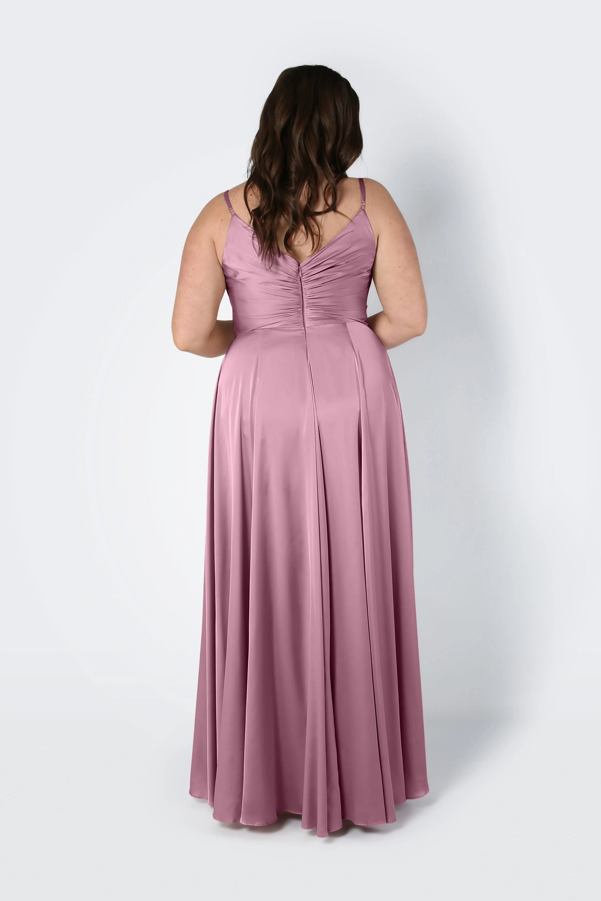 A woman with long brown hair faces away in the Isla Bridesmaid Dress by TH&TH, a rose pink satin gown with adjustable straps, gathered back, pleated bodice, softly flared floor-length skirt, set against a plain light background.