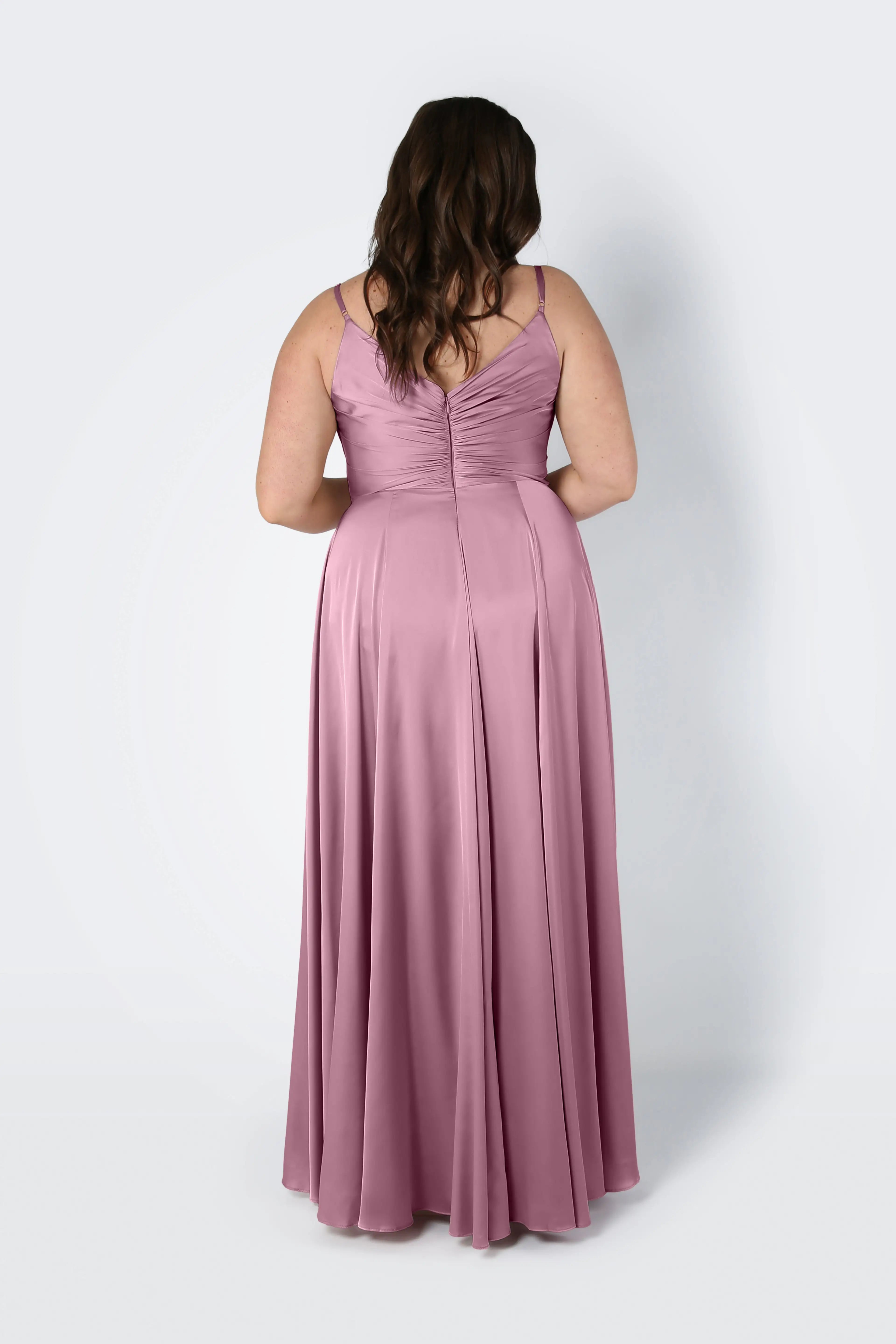 A woman with long brown hair faces away in the Isla Bridesmaid Dress by TH&TH, a rose pink satin gown with adjustable straps, gathered back, pleated bodice, softly flared floor-length skirt, set against a plain light background.