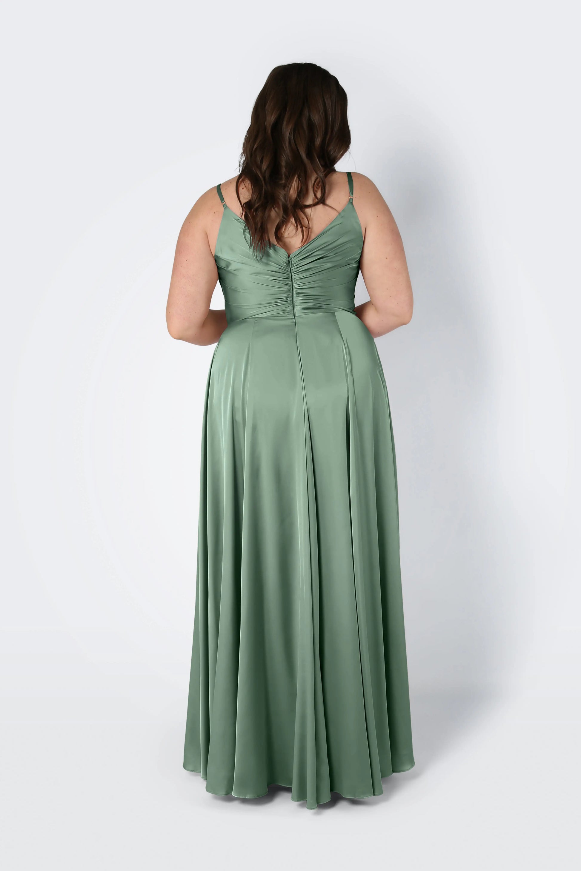 A woman with wavy brown hair, seen from behind, wears the TH&TH Isla Bridesmaid Dress in sage green satin—a long dress with thin straps and a fitted pleated bodice—standing against a plain light background.
