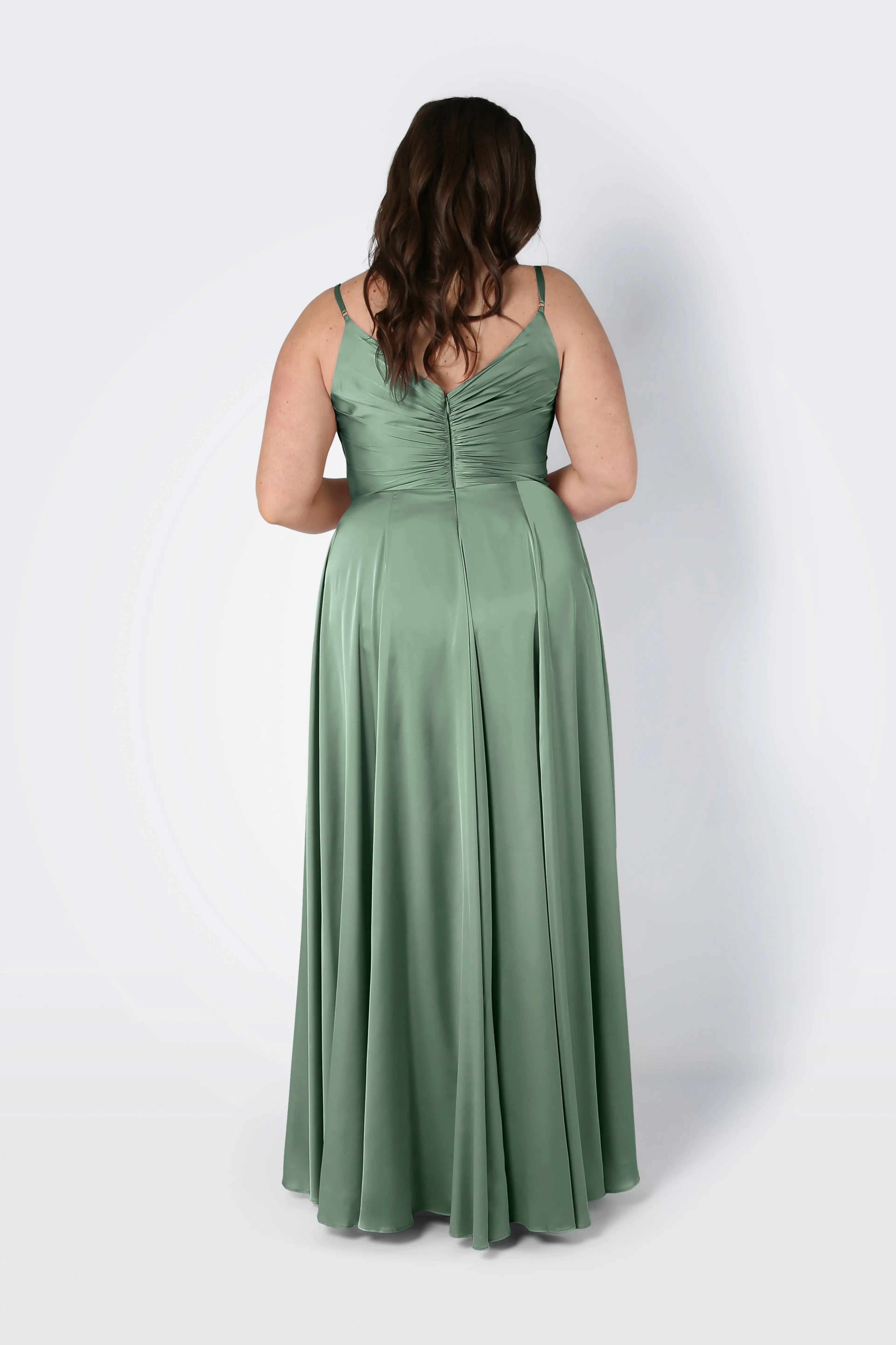 A woman with wavy brown hair, seen from behind, wears the TH&TH Isla Bridesmaid Dress in sage green satin—a long dress with thin straps and a fitted pleated bodice—standing against a plain light background.