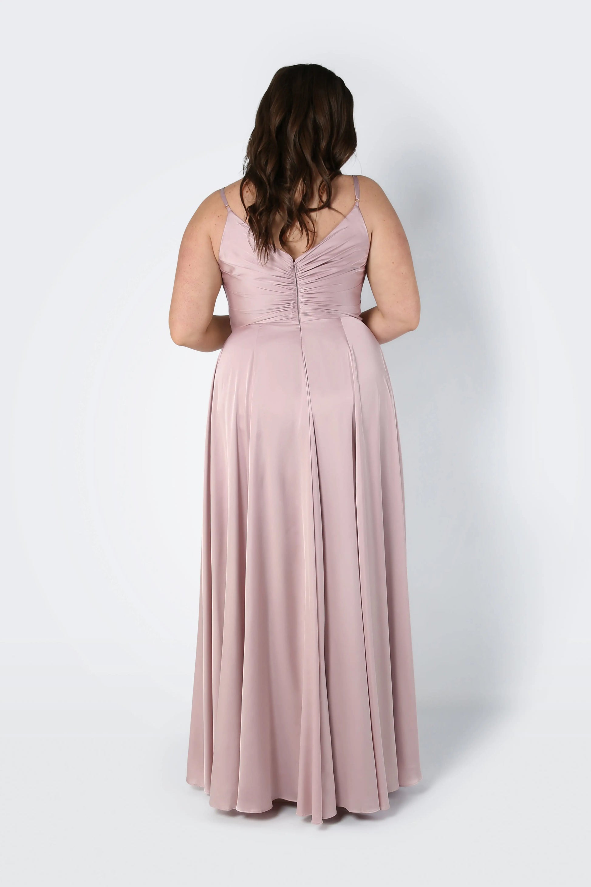 A woman with long wavy brown hair is seen from the back wearing the TH&TH Isla Bridesmaid Dress in Smoked Blush Satin, a sleeveless floor-length dress with a pleated fitted bodice and flowing skirt, against a plain light background.