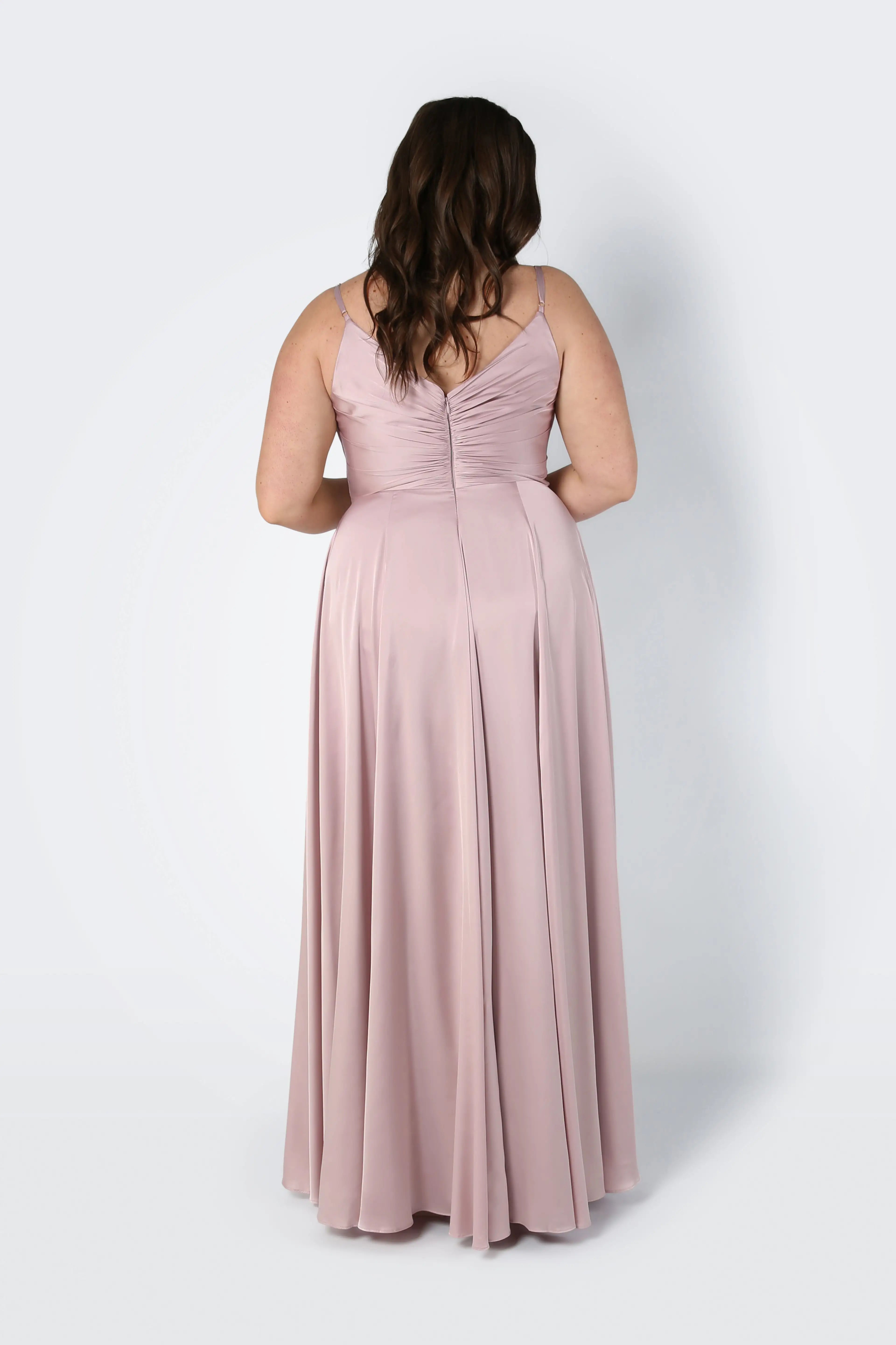 A woman with long wavy brown hair is seen from the back wearing the TH&TH Isla Bridesmaid Dress in Smoked Blush Satin, a sleeveless floor-length dress with a pleated fitted bodice and flowing skirt, against a plain light background.