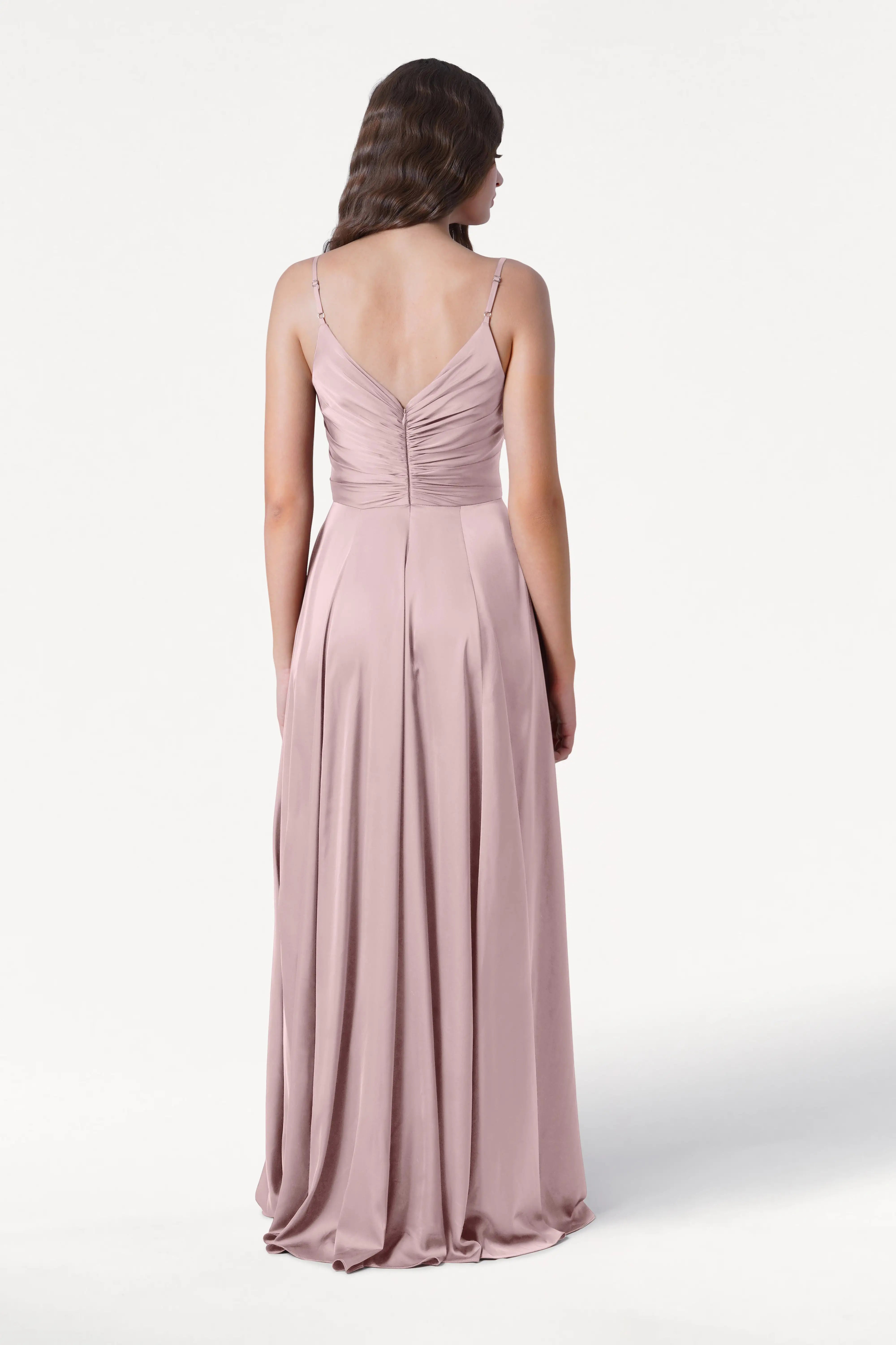 A woman wearing the TH&TH Isla Bridesmaid Dress in Smoked Blush Satin—with a pleated bodice, thin straps, and a ruched V-back—stands against a plain white background, facing away from the camera.