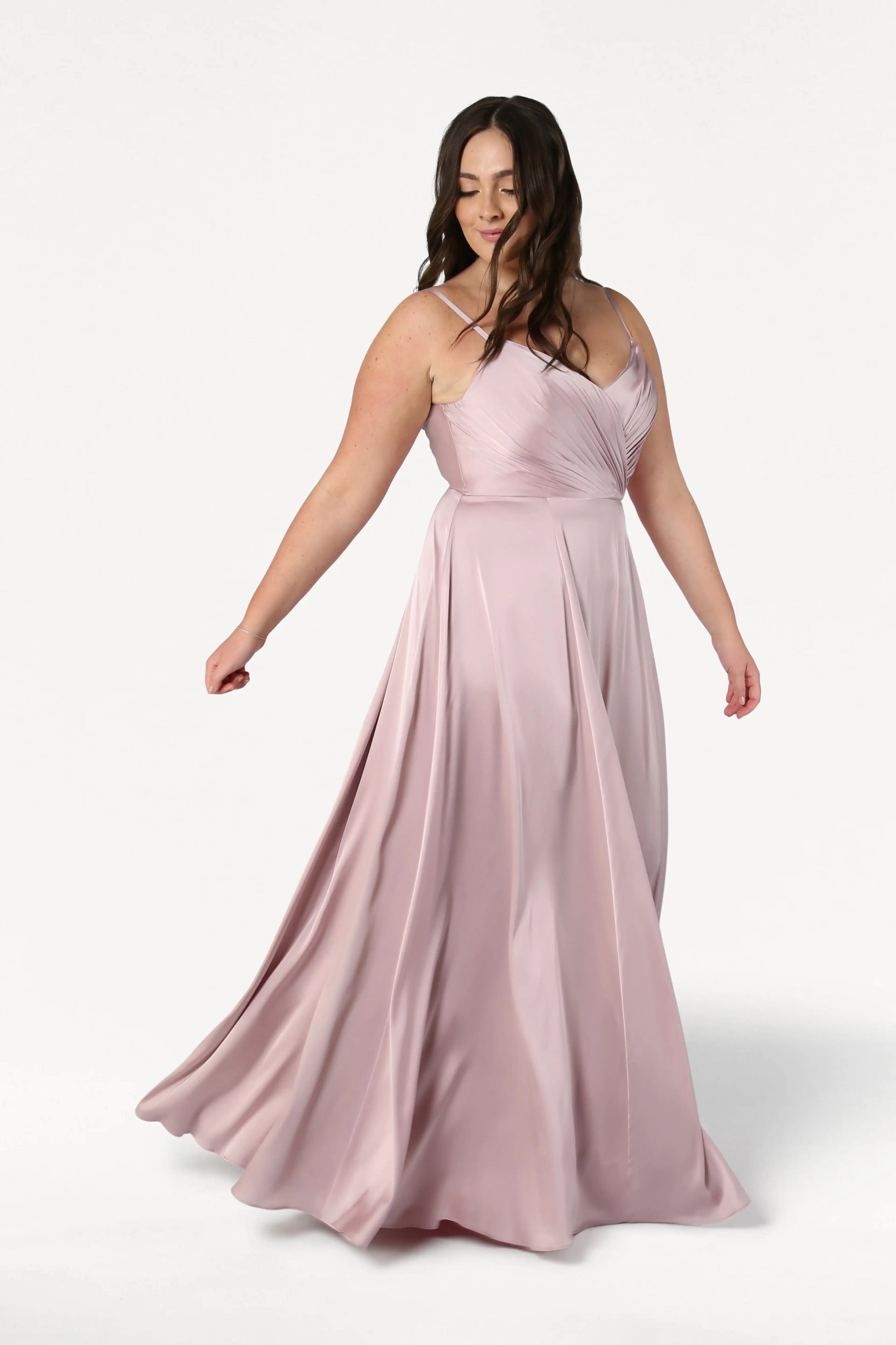 A woman with long brown hair twirls in the Isla Bridesmaid Dress in Smoked Blush Satin by TH&TH, featuring a pleated bodice and thin straps. The satin fabric creates gentle movement against a plain white background.