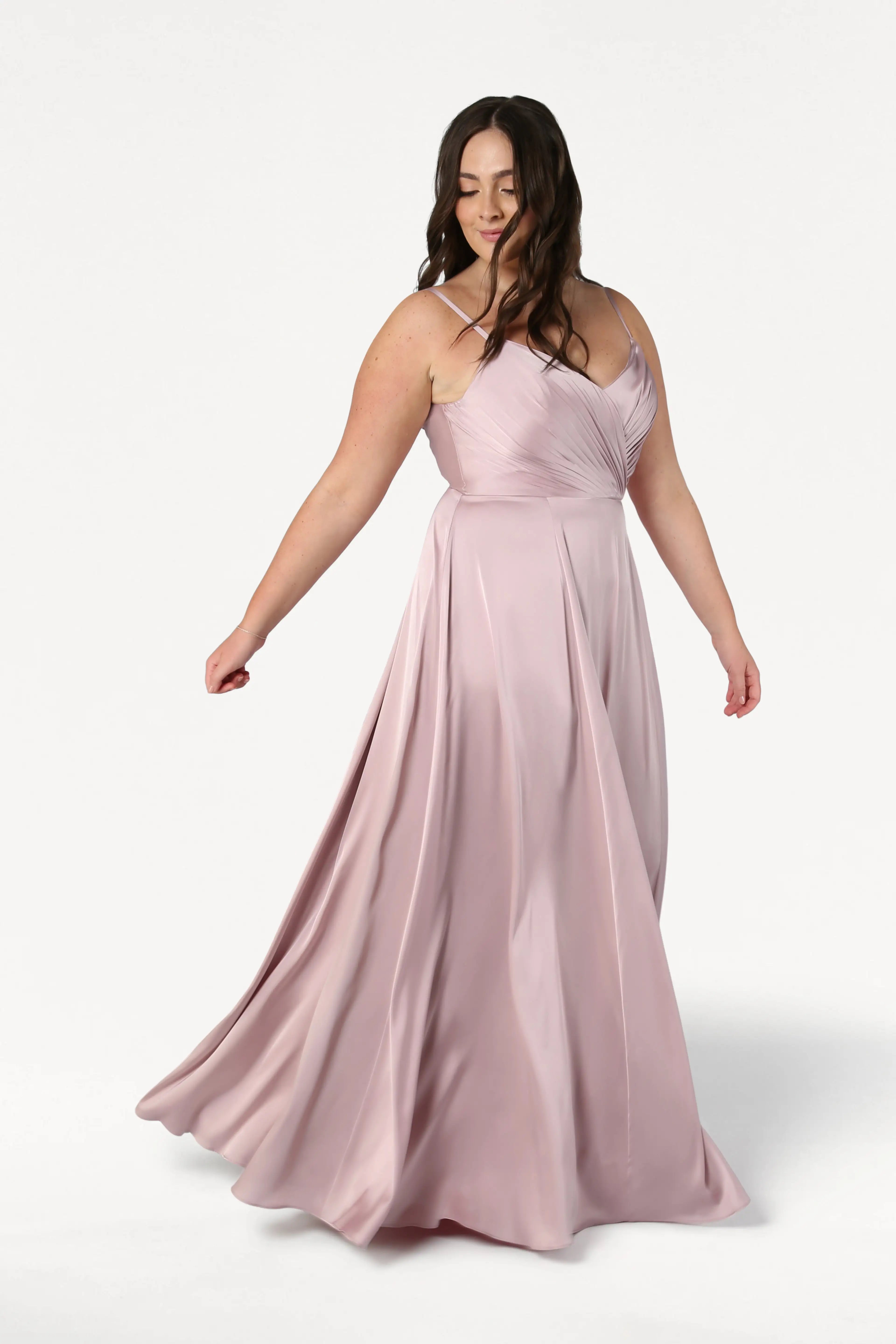 A woman with long brown hair twirls in the Isla Bridesmaid Dress in Smoked Blush Satin by TH&TH, featuring a pleated bodice and thin straps. The satin fabric creates gentle movement against a plain white background.