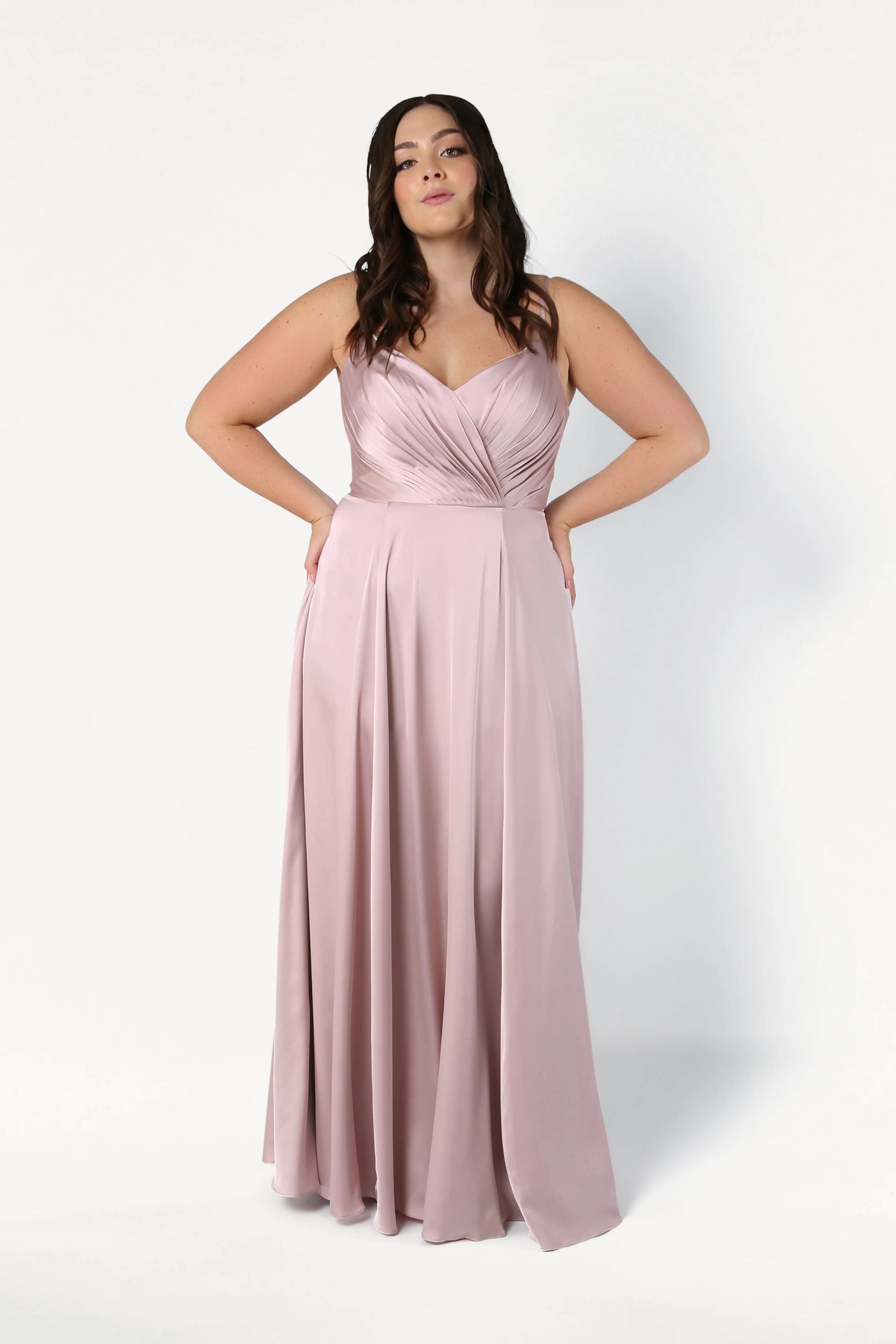 A woman with long, wavy brown hair wears the TH&TH Isla Bridesmaid Dress in Smoked Blush Satin, a sleeveless, floor-length gown with a pleated bodice and flowy skirt. She stands against a plain white background with hands on her hips.