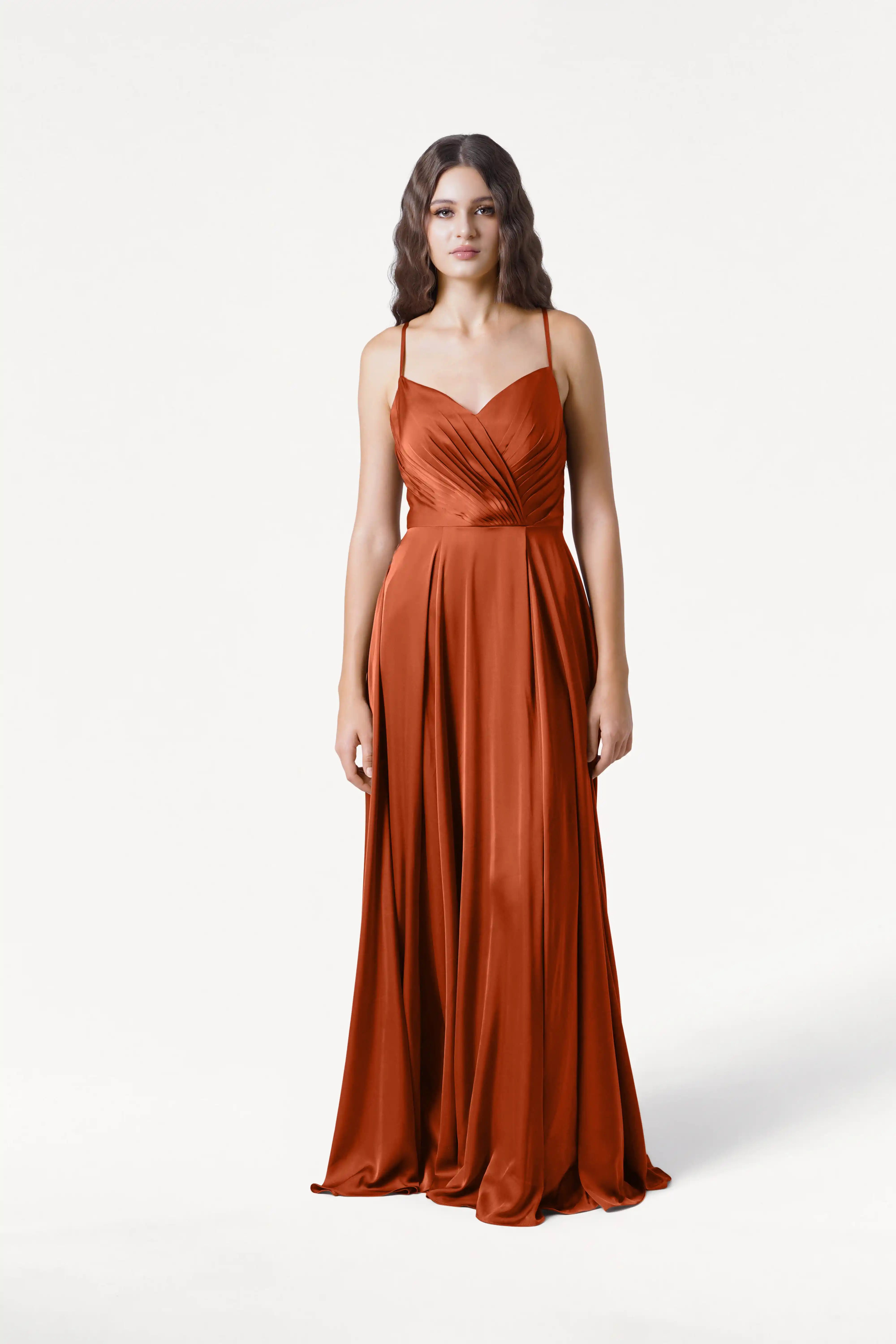 A woman with softly waved hair stands against a white background wearing the TH&TH Isla Bridesmaid Dress in Terracotta Satin, featuring a long silhouette, thin straps, and a draped, pleated bodice.
