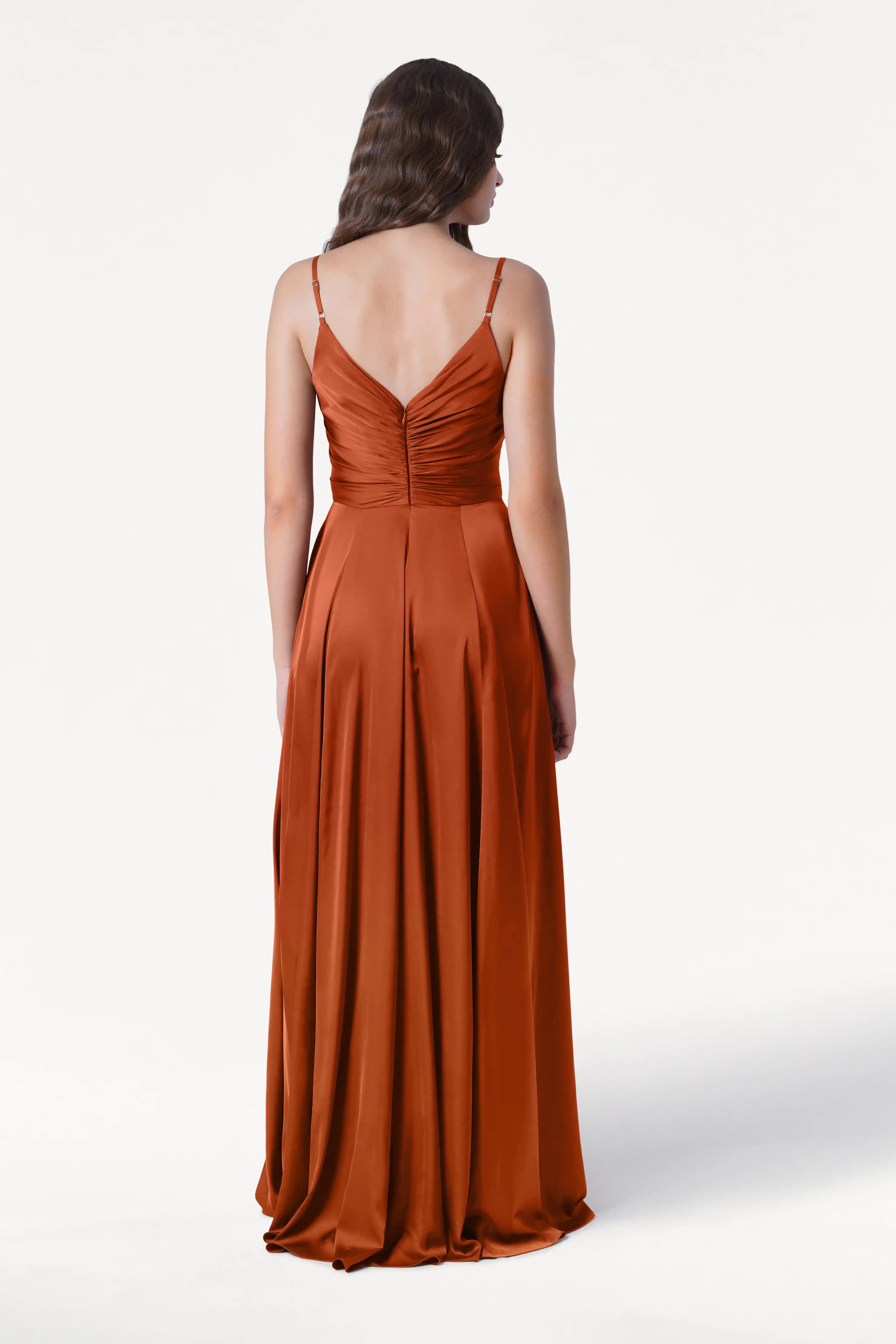 A woman faces away, wearing the TH&TH Isla Bridesmaid Dress in Terracotta Satin, featuring thin straps and a fitted, pleated bodice with gentle pleats flowing to the floor. The background is plain white.