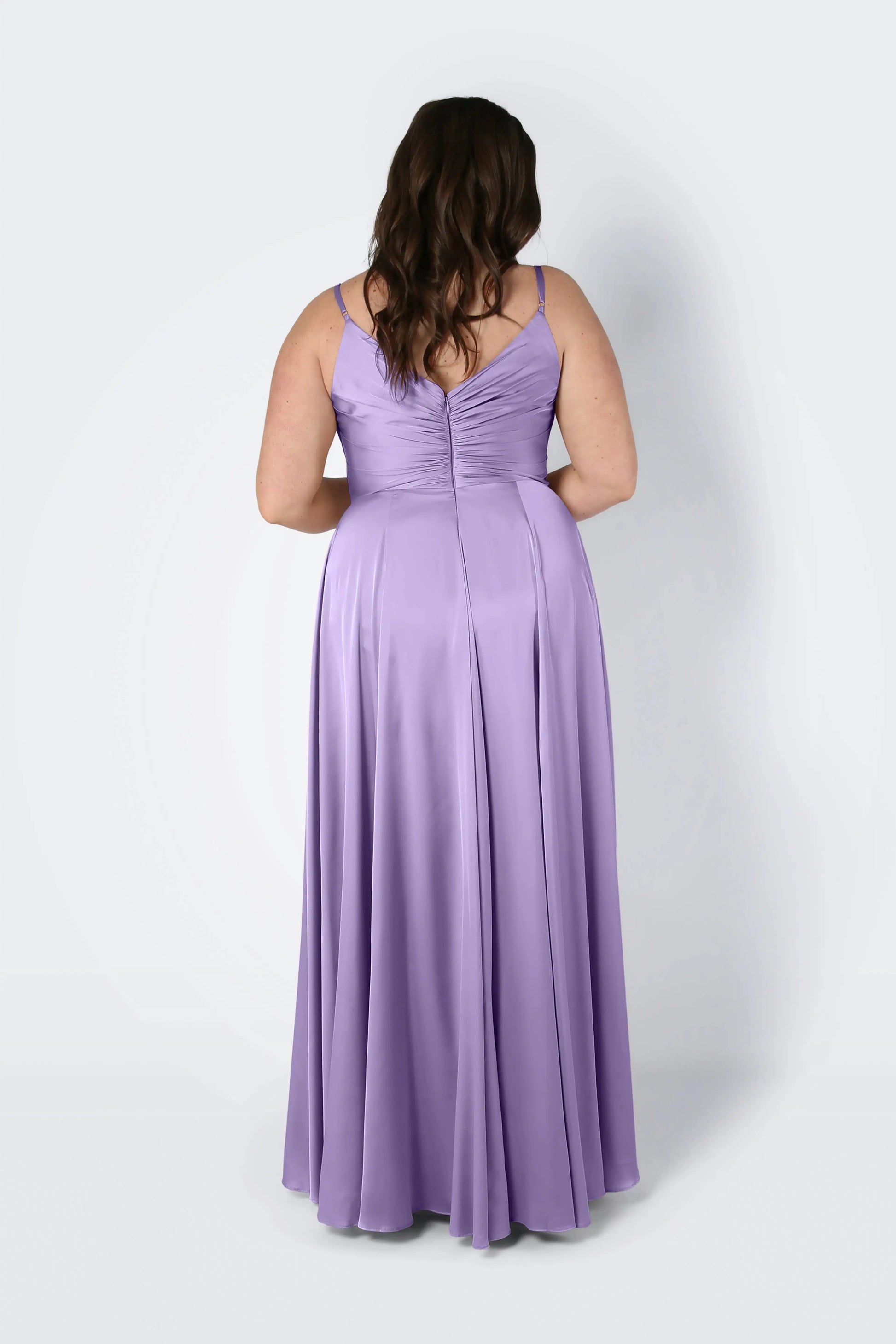 A woman with wavy brown hair faces away, wearing the TH&TH Isla Bridesmaid Dress in Violet Satin—a long, sleeveless gown with a pleated bodice and flowing skirt—set against a plain light background.