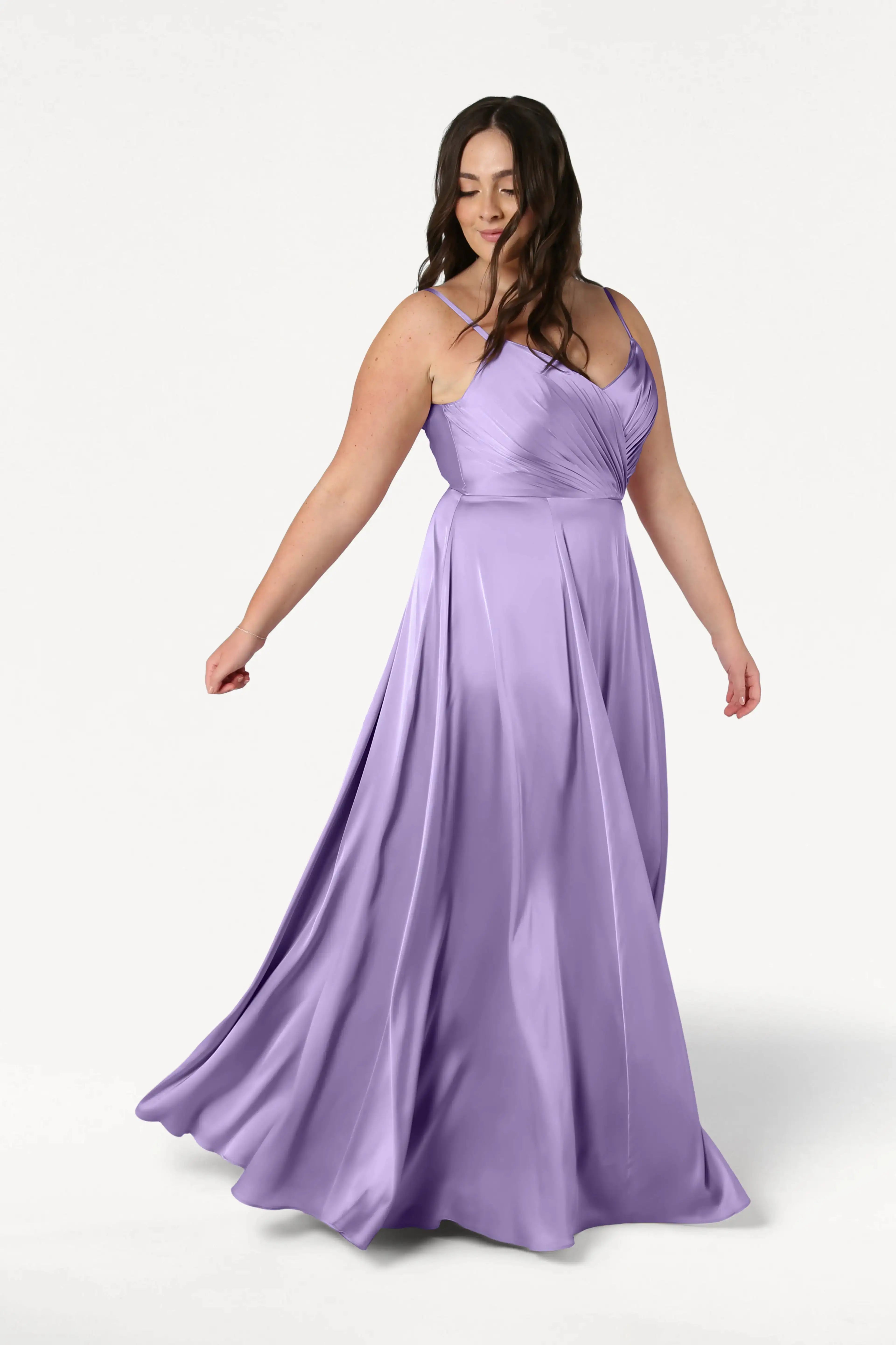 A woman wearing the Isla Bridesmaid Dress in Violet Satin by TH&TH stands against a plain white background, gently smiling as she holds out part of her long, pleated gown.