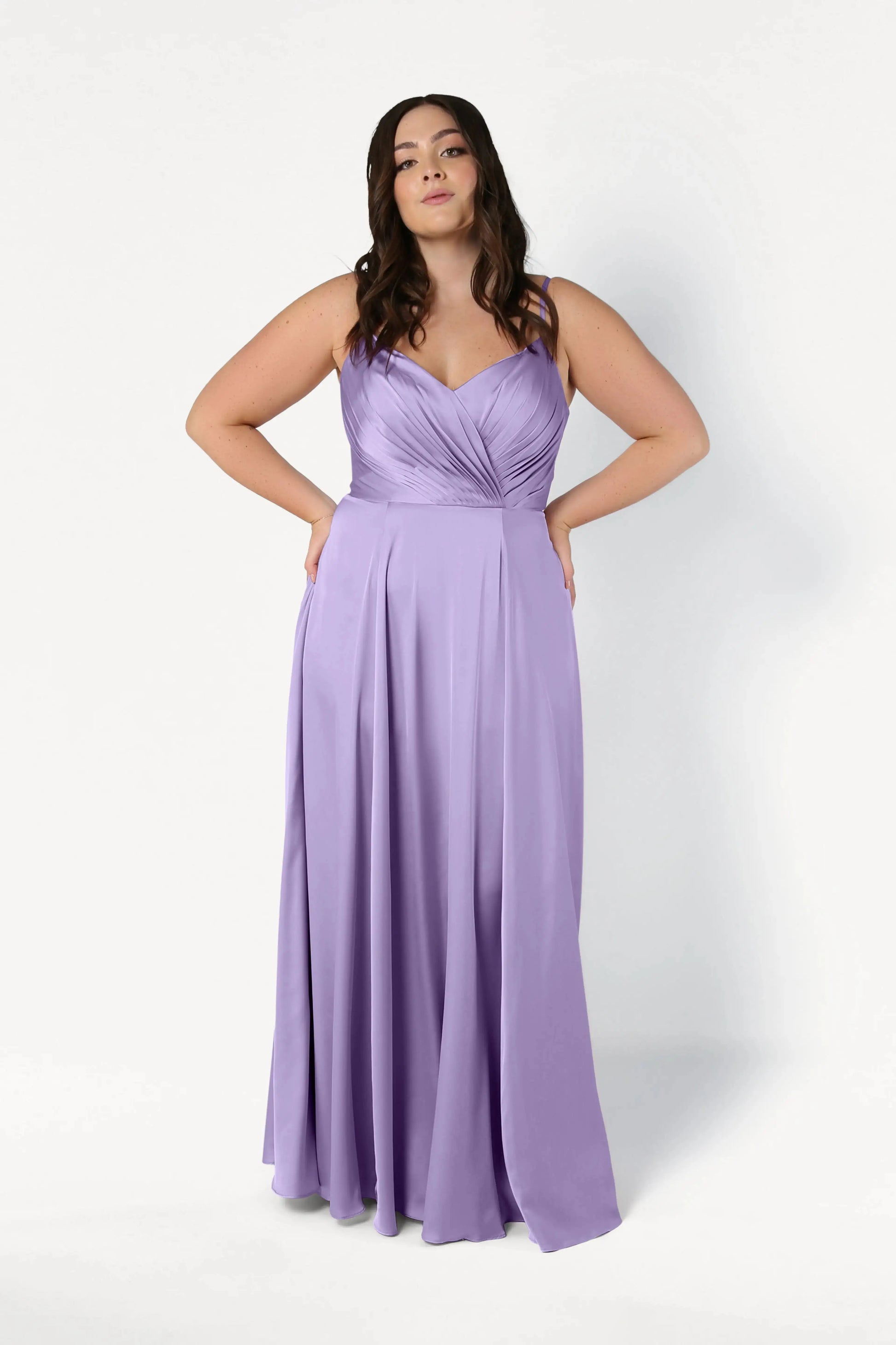 A woman stands confidently in the TH&TH Isla Bridesmaid Dress in Violet Satin, featuring a pleated bodice and flowing skirt, against a plain white background.