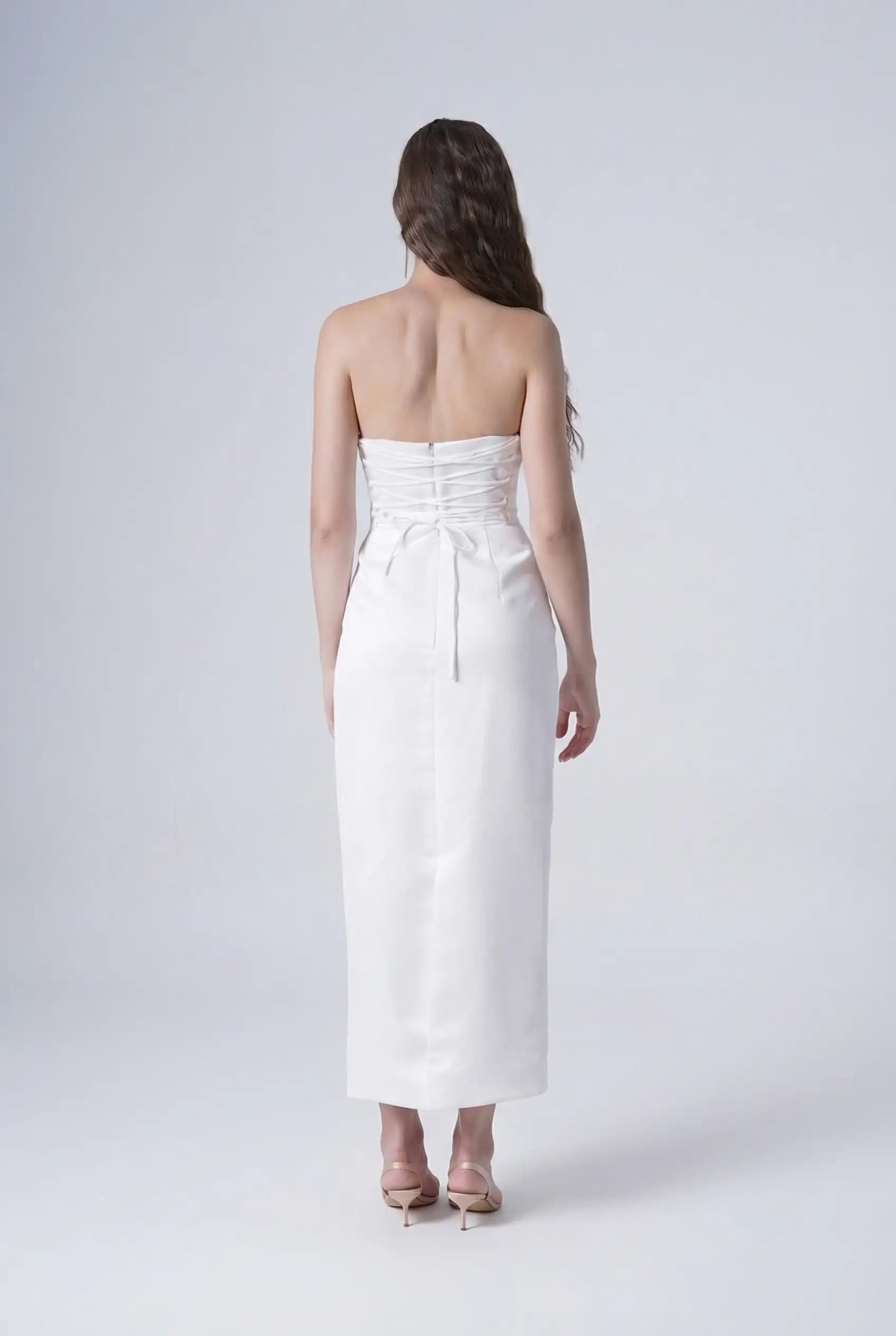 A woman with long wavy hair faces away, wearing the TH&TH Ivy Bridal Dress in Ivory and nude heels, set against a plain light background.