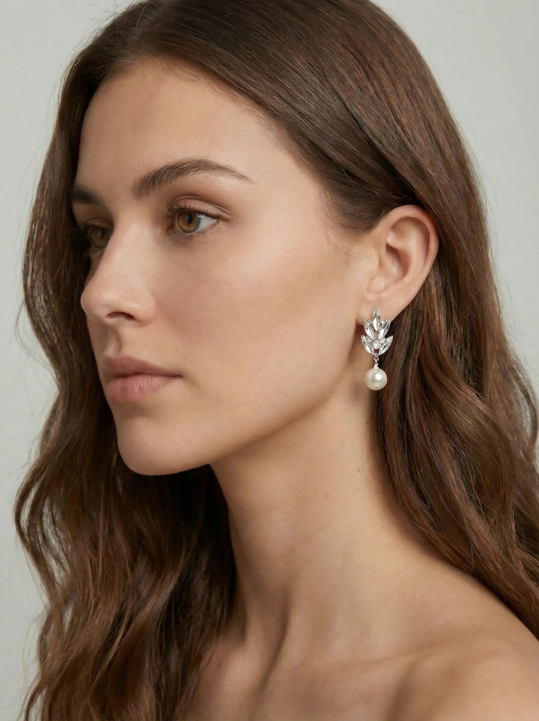 A woman with long brown hair wears TH&TH Laurel Crystal Drop Earrings—leaf-shaped and adorned with crystals and pearls—for a bridal look. She has a neutral expression, bare shoulders, and is set against a plain, light background.