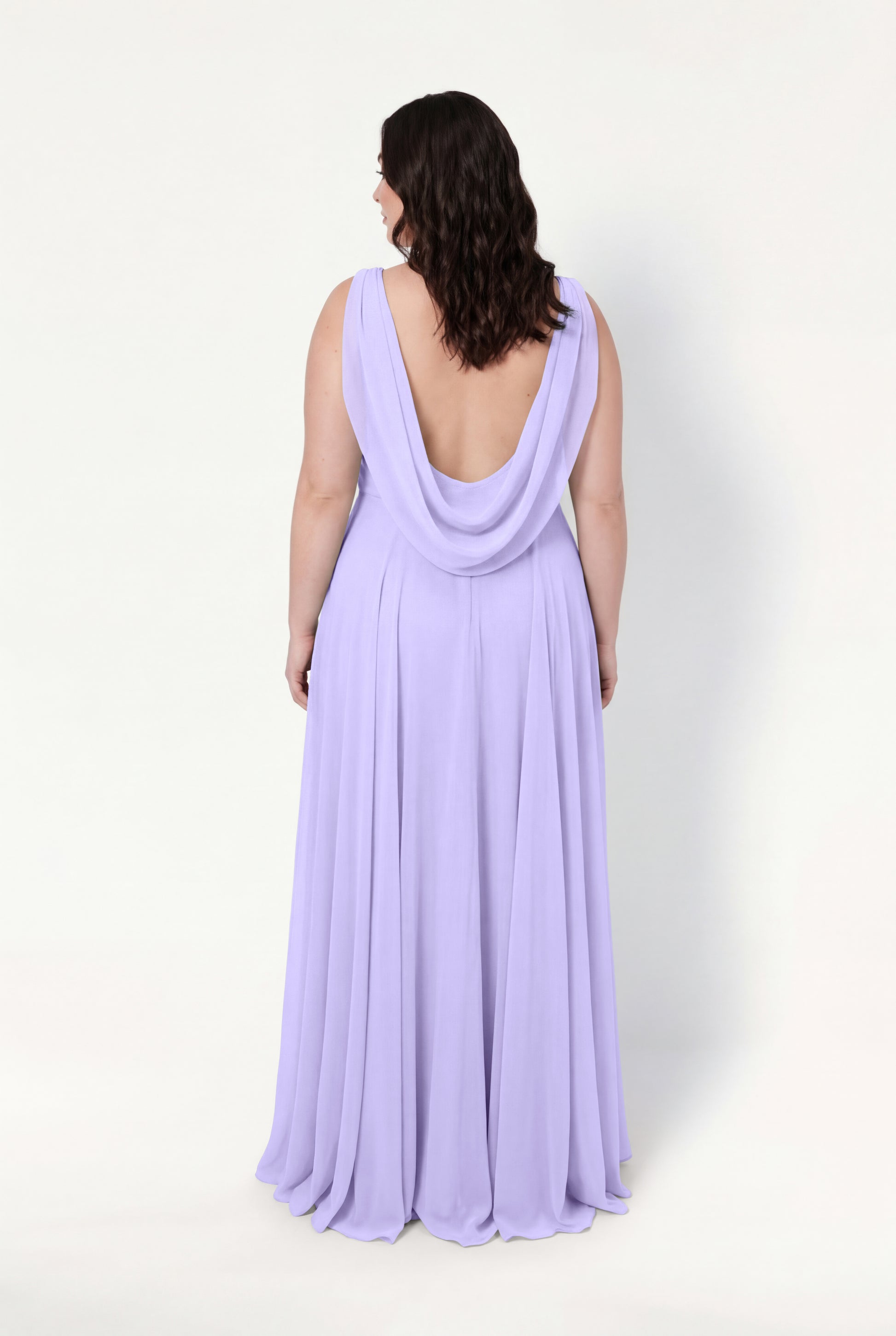A woman with wavy brown hair models the TH&TH Athena Bridesmaid Dress in Lilac Mist chiffon, a floor-length, sleeveless gown with a draped low cowl back, standing against a plain white background and facing away from the camera.