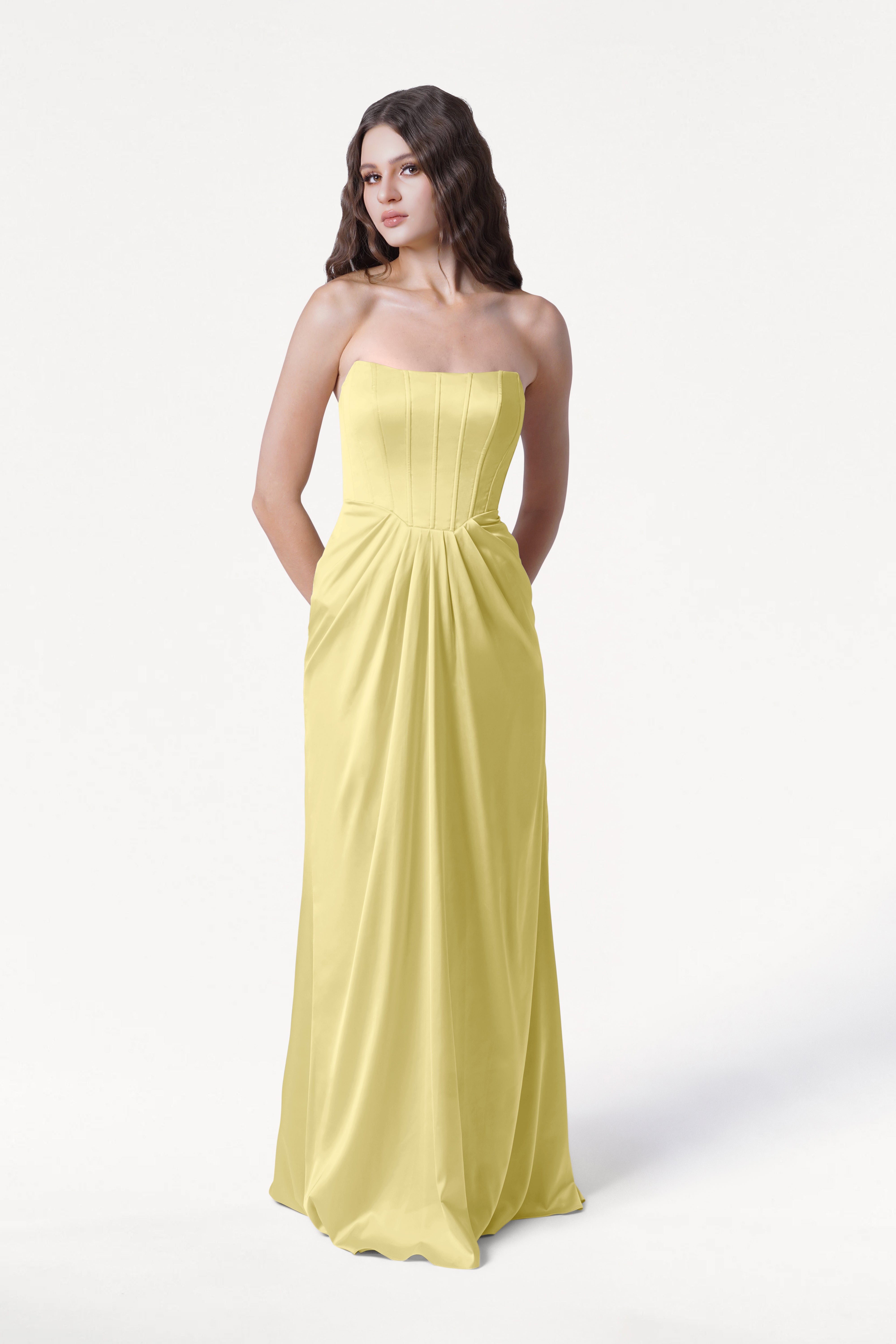 A woman with long brown hair wears the TH&TH Selena Bridesmaid Dress in Limoncello Satin, a strapless, floor-length gown with side pockets, standing against a plain white background.