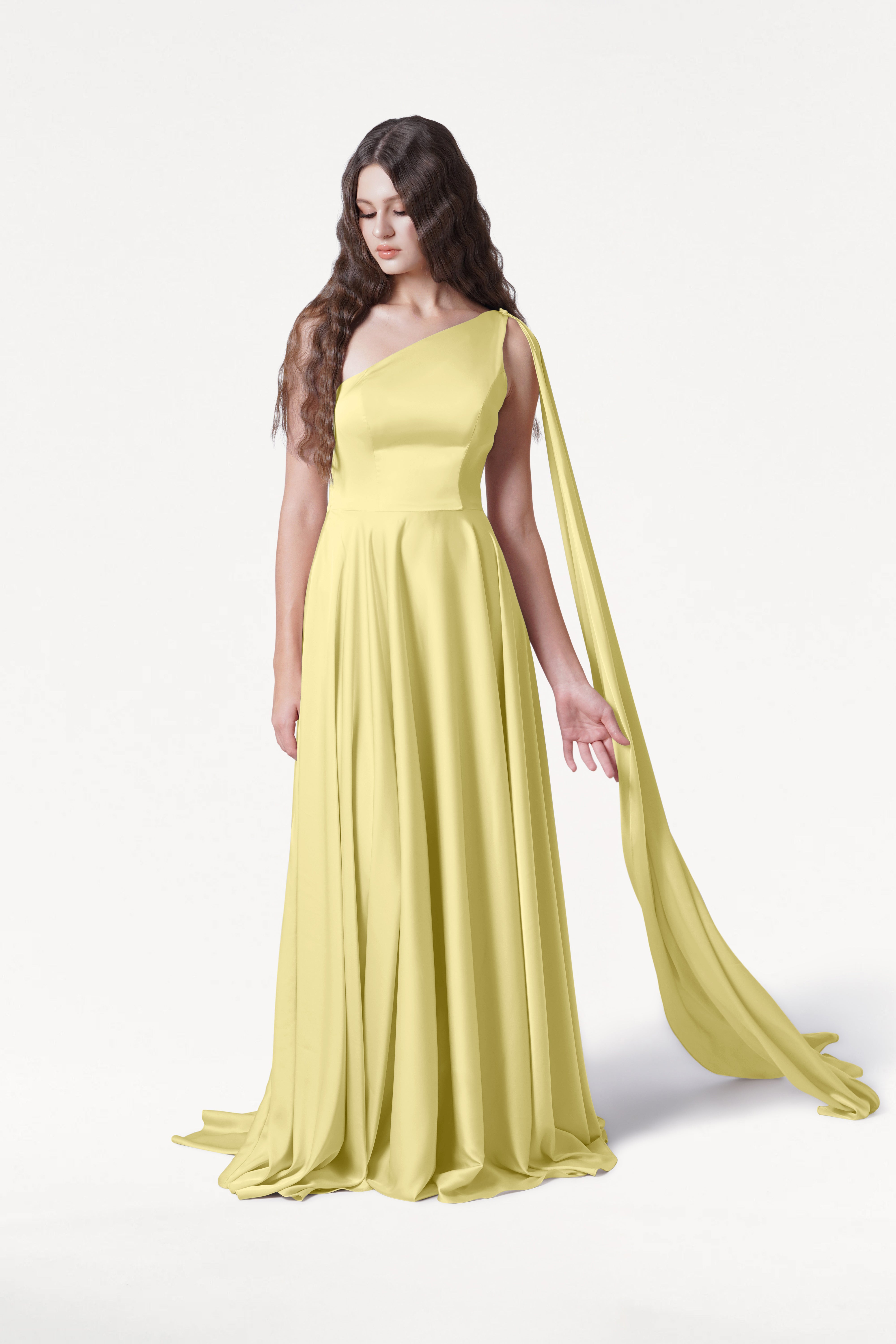 A woman with long, wavy brown hair wears the TH&TH Lyra Bridesmaid Dress in Limoncello Satin, a size-inclusive, elegant one-shoulder gown with a draped sash, standing against a white background and looking down gracefully.