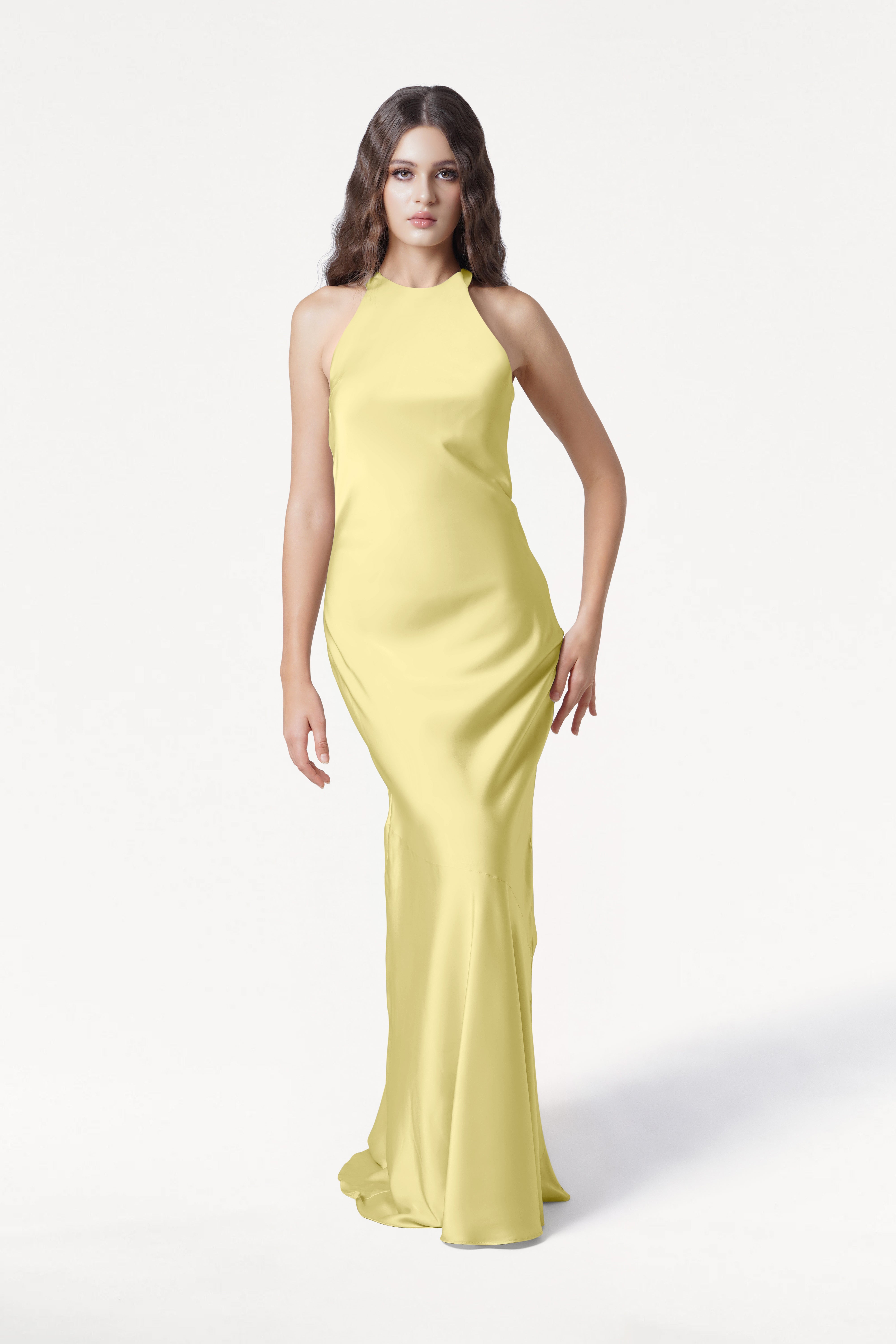 A woman with long brown hair in loose waves and a neutral expression wears the TH&TH Mira Bridesmaid Dress in Limoncello Satin, a sleeveless, floor-length halterneck gown, standing against a white background.
