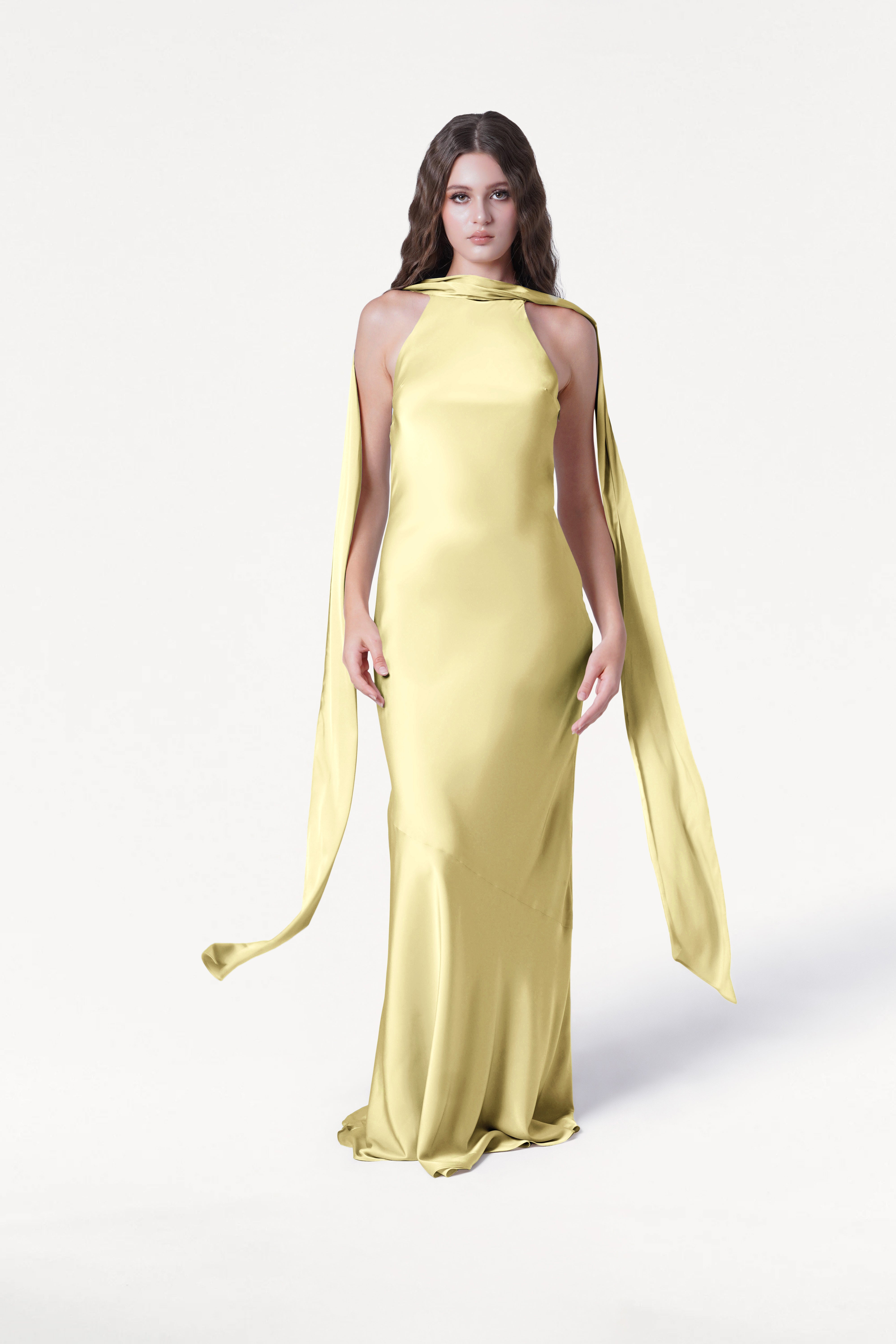 A woman with long, wavy brown hair and a neutral expression wears the TH&TH Mira Bridesmaid Dress in Limoncello Satin—floor-length, bias-cut, featuring draped scarf-like fabric—while standing against a plain white background.