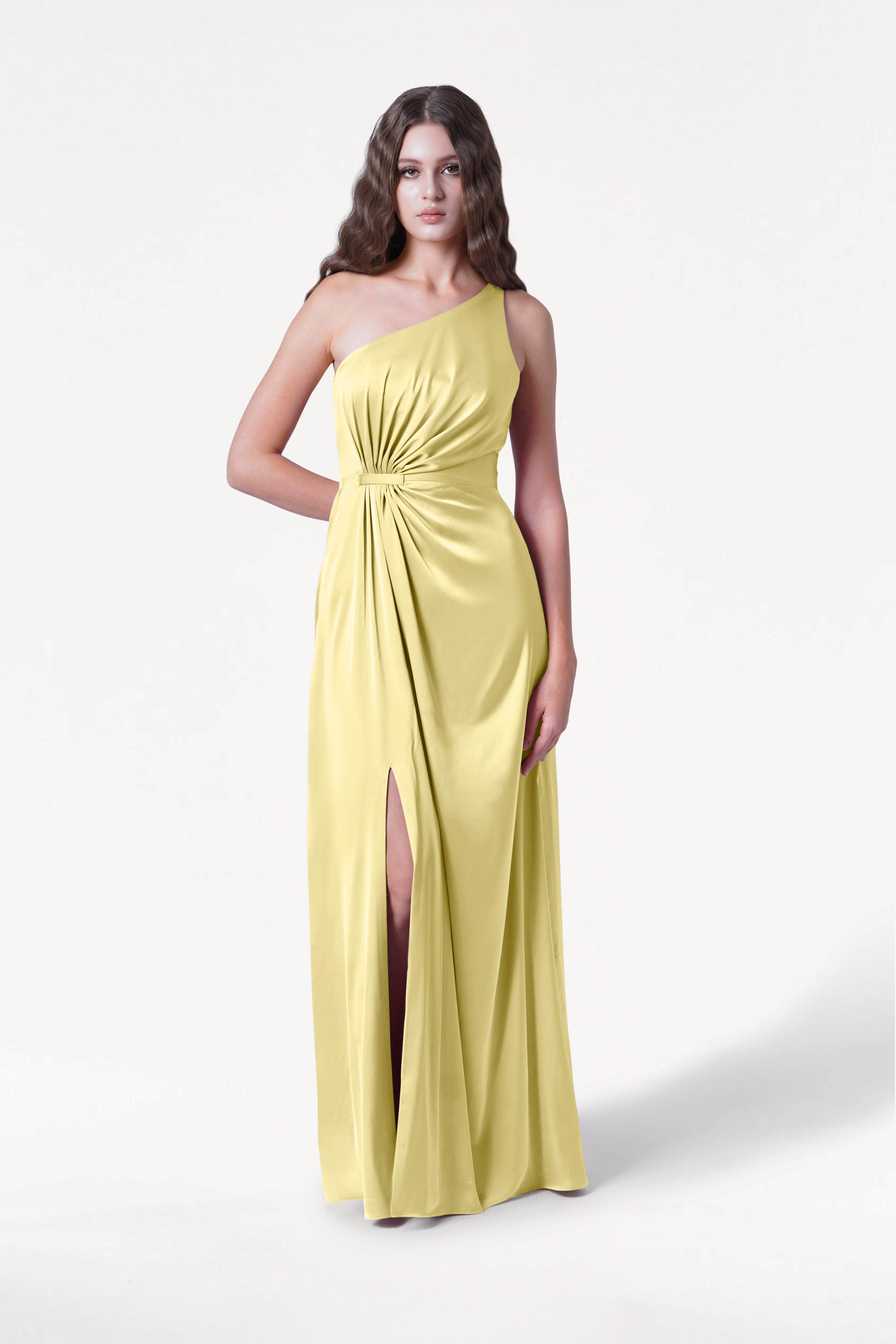 A woman with long, wavy brown hair and a neutral expression models the TH&TH Greta Bridesmaid Dress in Limoncello Satin—a long, one-shoulder gown with Grecian draping, a gathered knot waist, and high slit—against a white background.