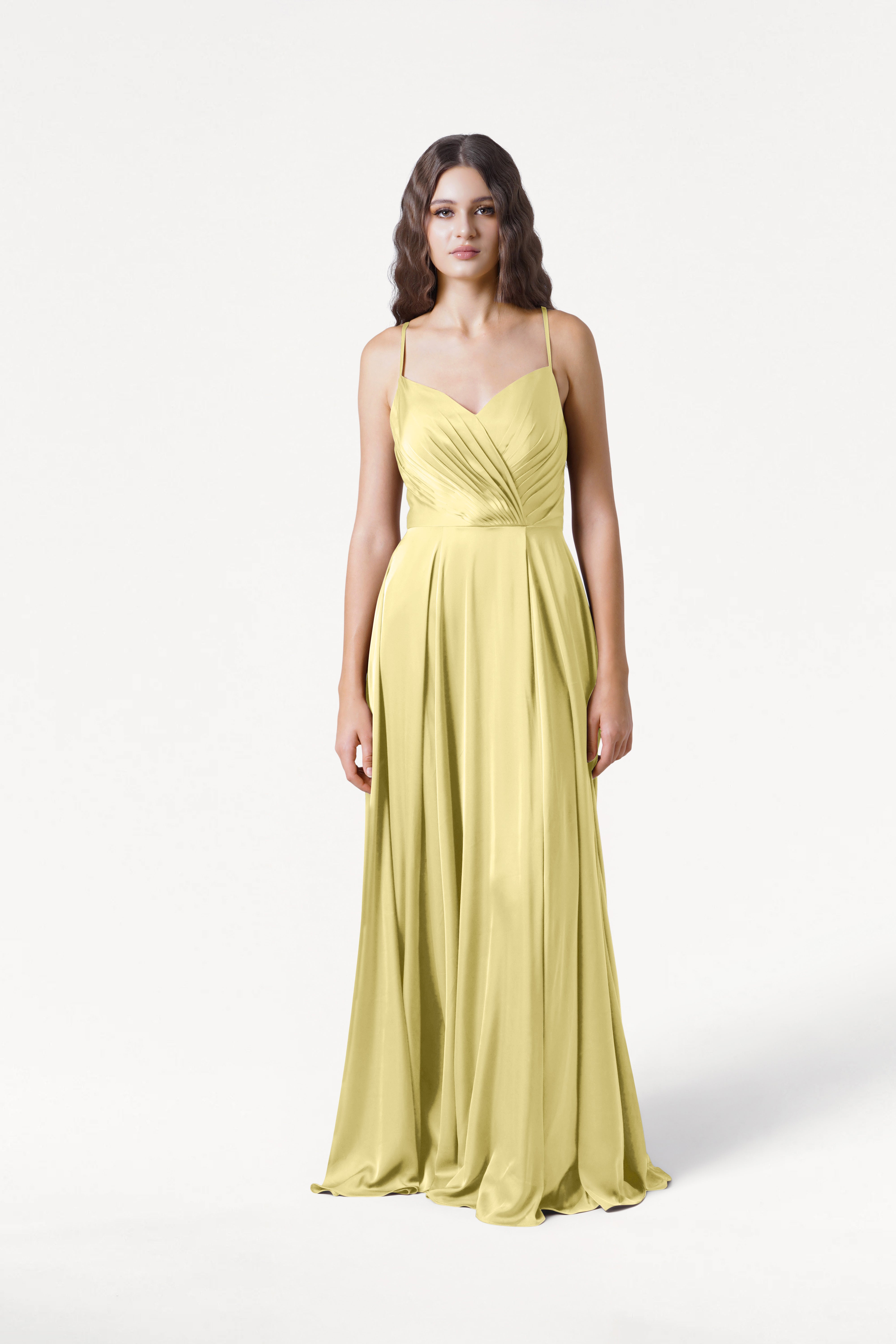 A woman with long, wavy hair and a neutral expression stands against a white background wearing the TH&TH Isla Bridesmaid Dress in Limoncello Satin, a long yellow gown with thin straps and a fitted, pleated bodice.