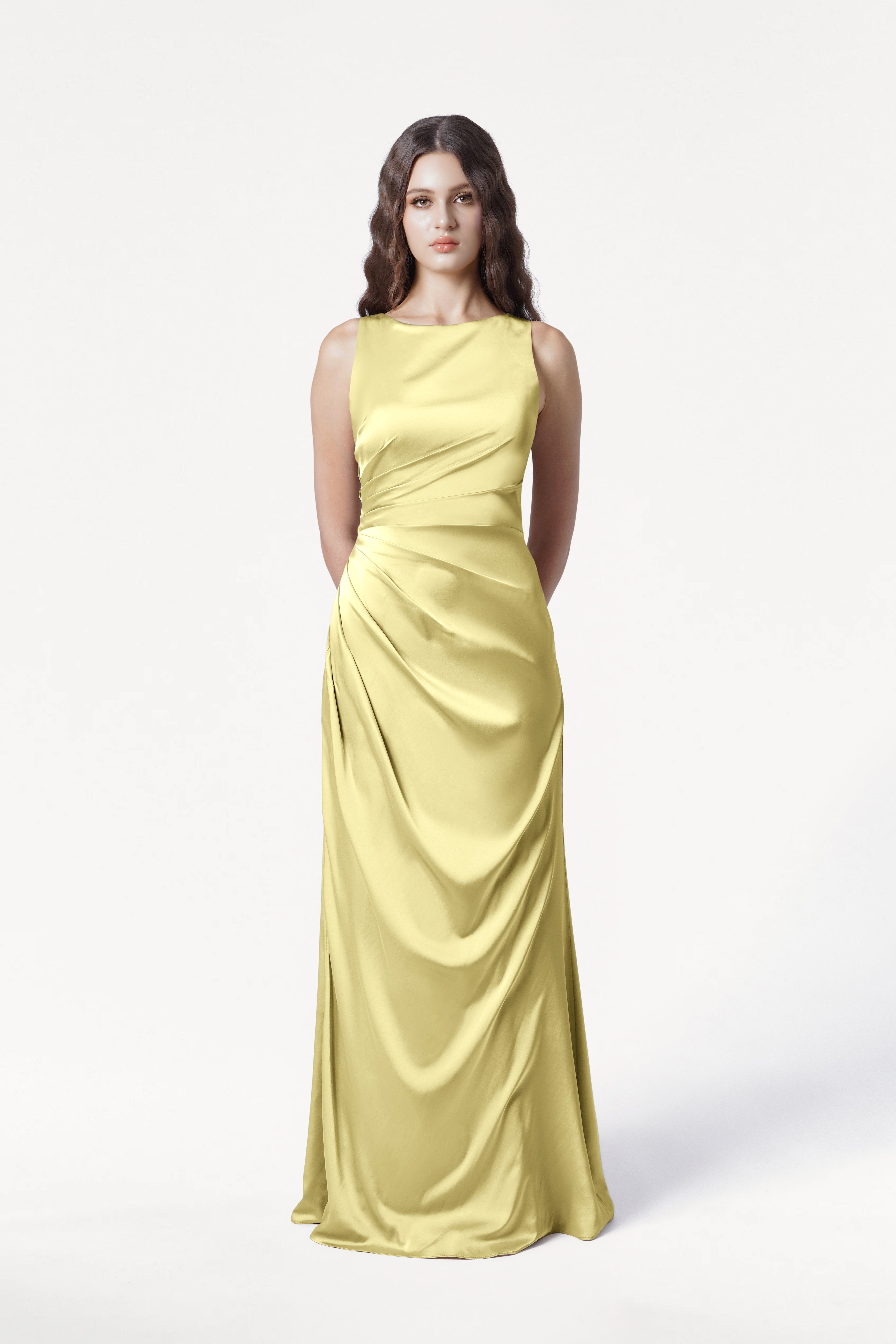 A woman with long, wavy brown hair models the TH&TH Nala Bridesmaid Dress in Limoncello Satin, a size-inclusive, floor-length gown with Grecian-inspired pleating and gathered draping, against a plain white background.