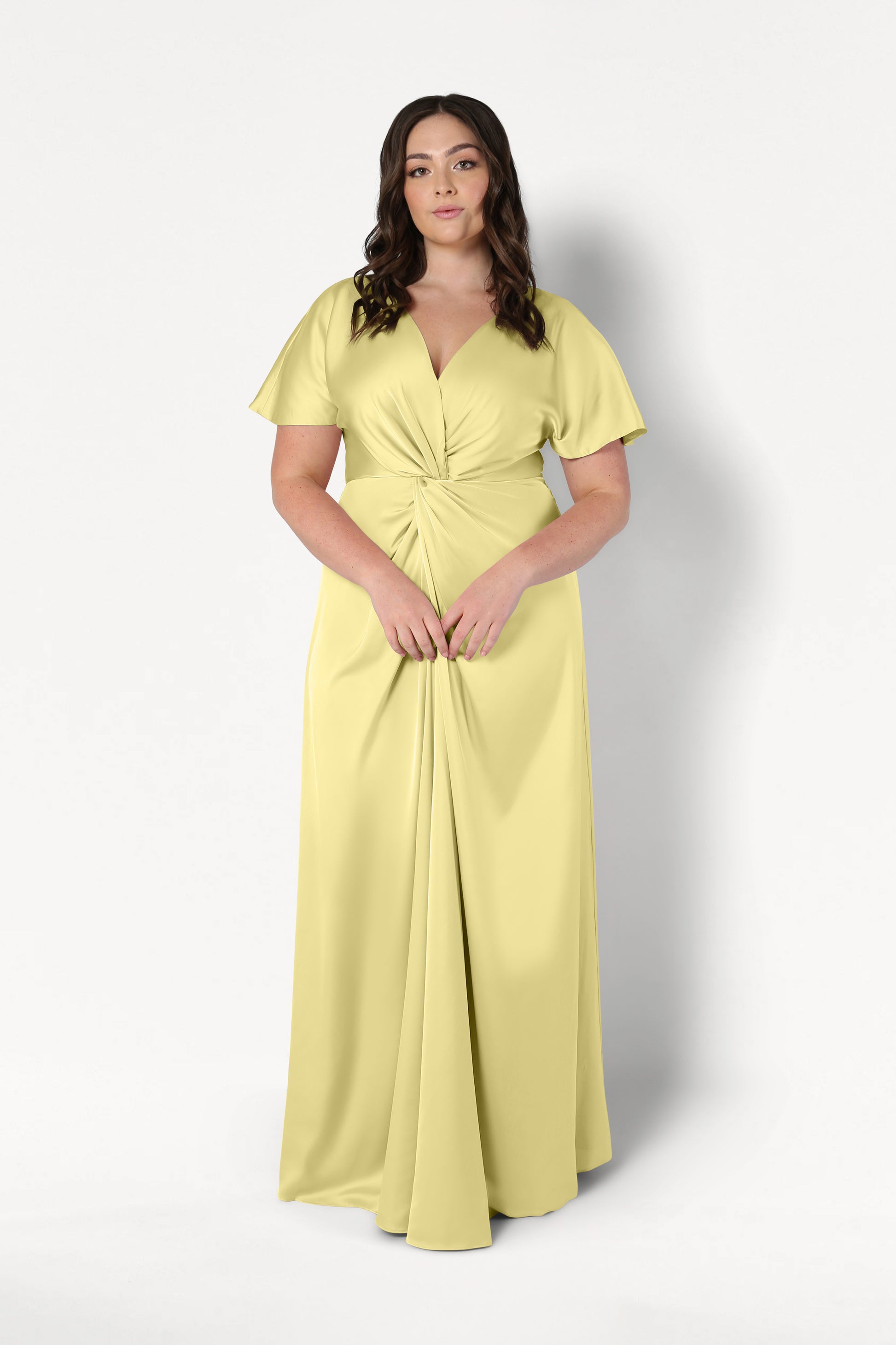 A woman with long dark wavy hair wears the TH&TH Camilla Bridesmaid Dress in Limoncello Satin, a floor-length gown with short sleeves and a twist-knot bodice, standing against a plain white background.
