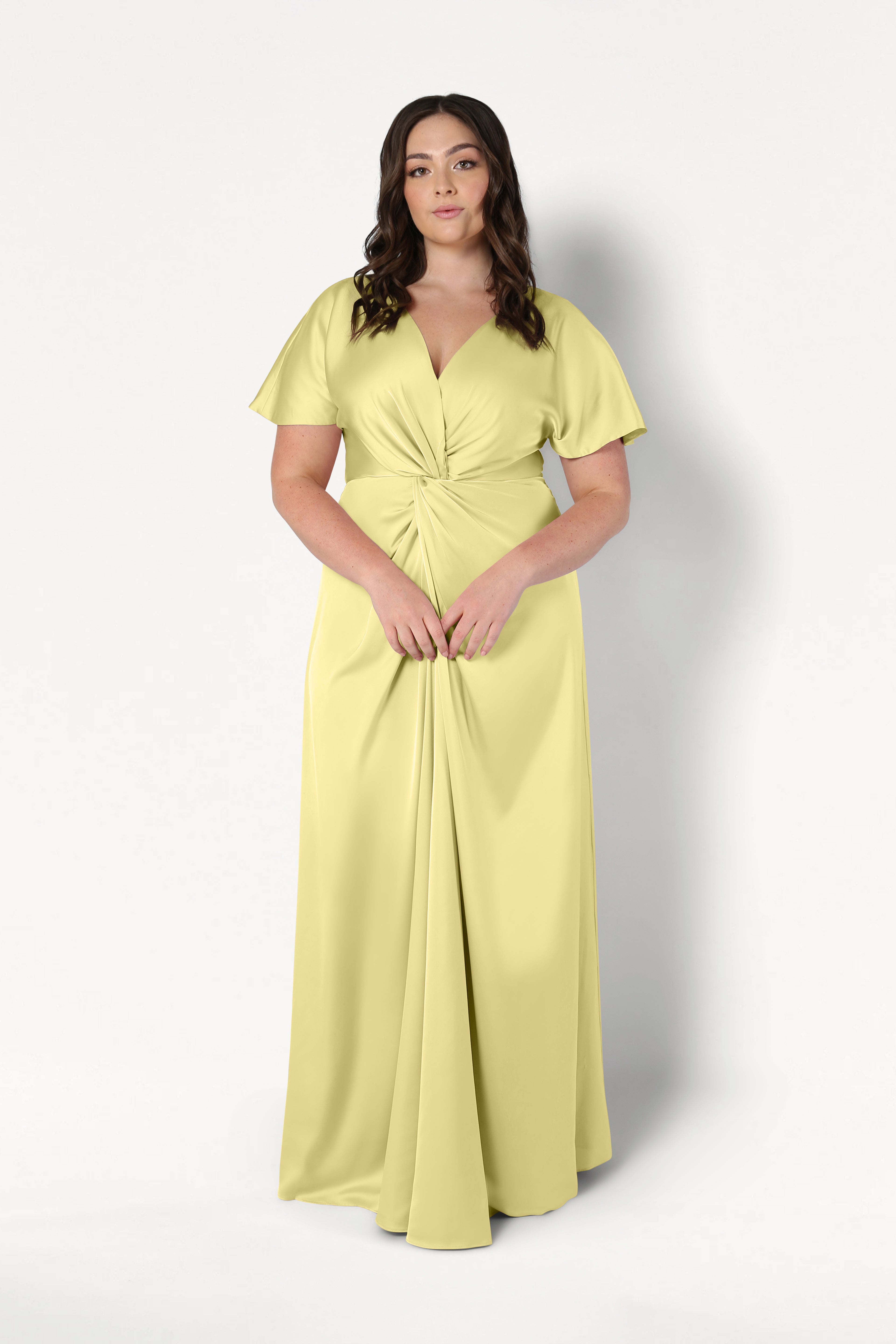 A woman with long dark wavy hair wears the TH&TH Camilla Bridesmaid Dress in Limoncello Satin, a floor-length gown with short sleeves and a twist-knot bodice, standing against a plain white background.