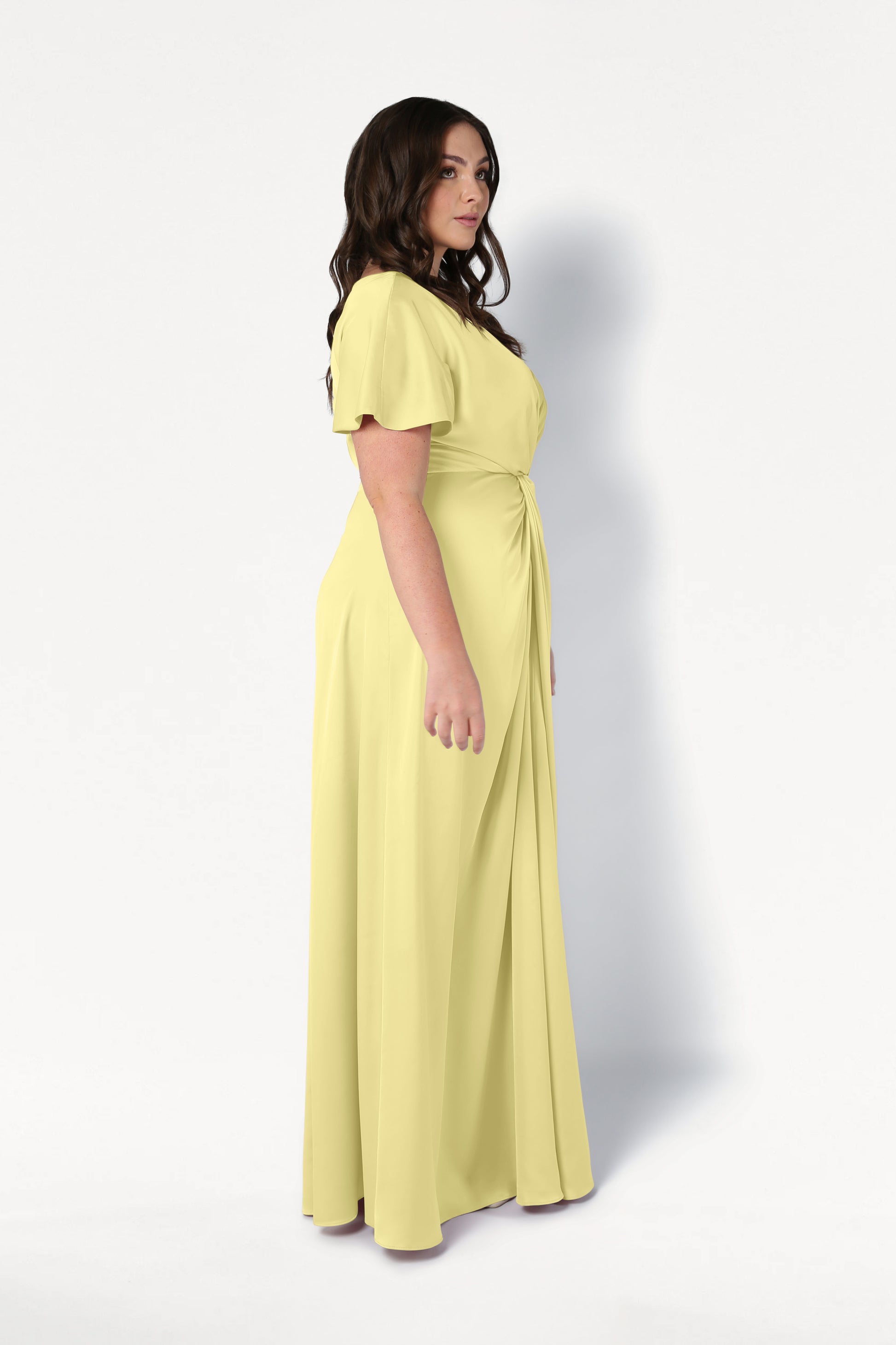 A woman with long brown hair stands in profile against a plain white background, wearing the TH&TH Camilla Bridesmaid Dress in Limoncello Satin—a size-inclusive, floor-length gown with short sleeves and a gathered waist.