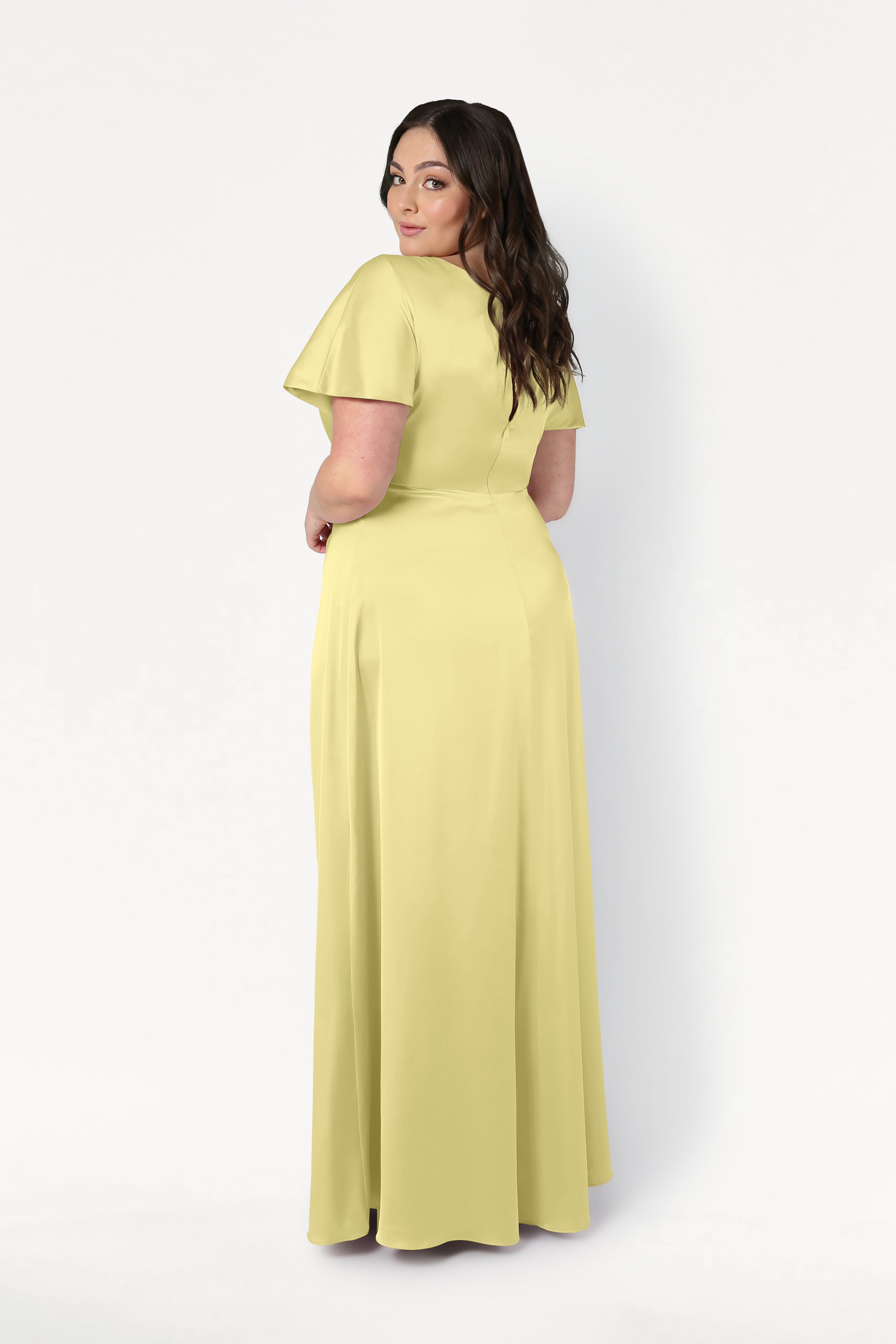 A woman with long, dark wavy hair stands looking over her shoulder in the TH&TH Camilla Bridesmaid Dress in Limoncello Satin, featuring a twist-knot bodice. The photo is set against a plain white background.