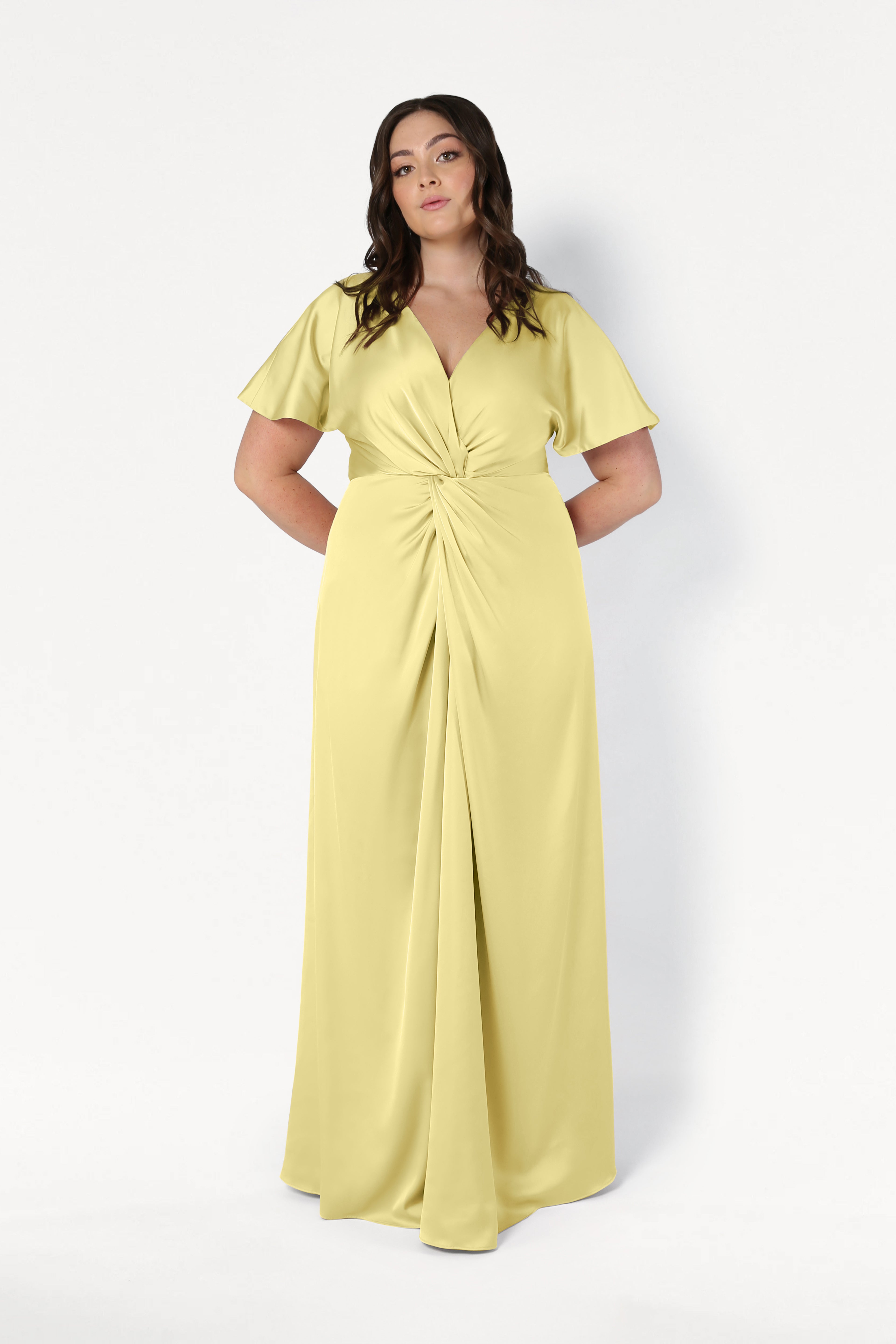 A woman with long wavy hair and a neutral expression poses against a white background wearing the TH&TH Camilla Bridesmaid Dress in Limoncello Satin, featuring short flutter sleeves, a V-neckline, gathered waist, and size-inclusive fit.