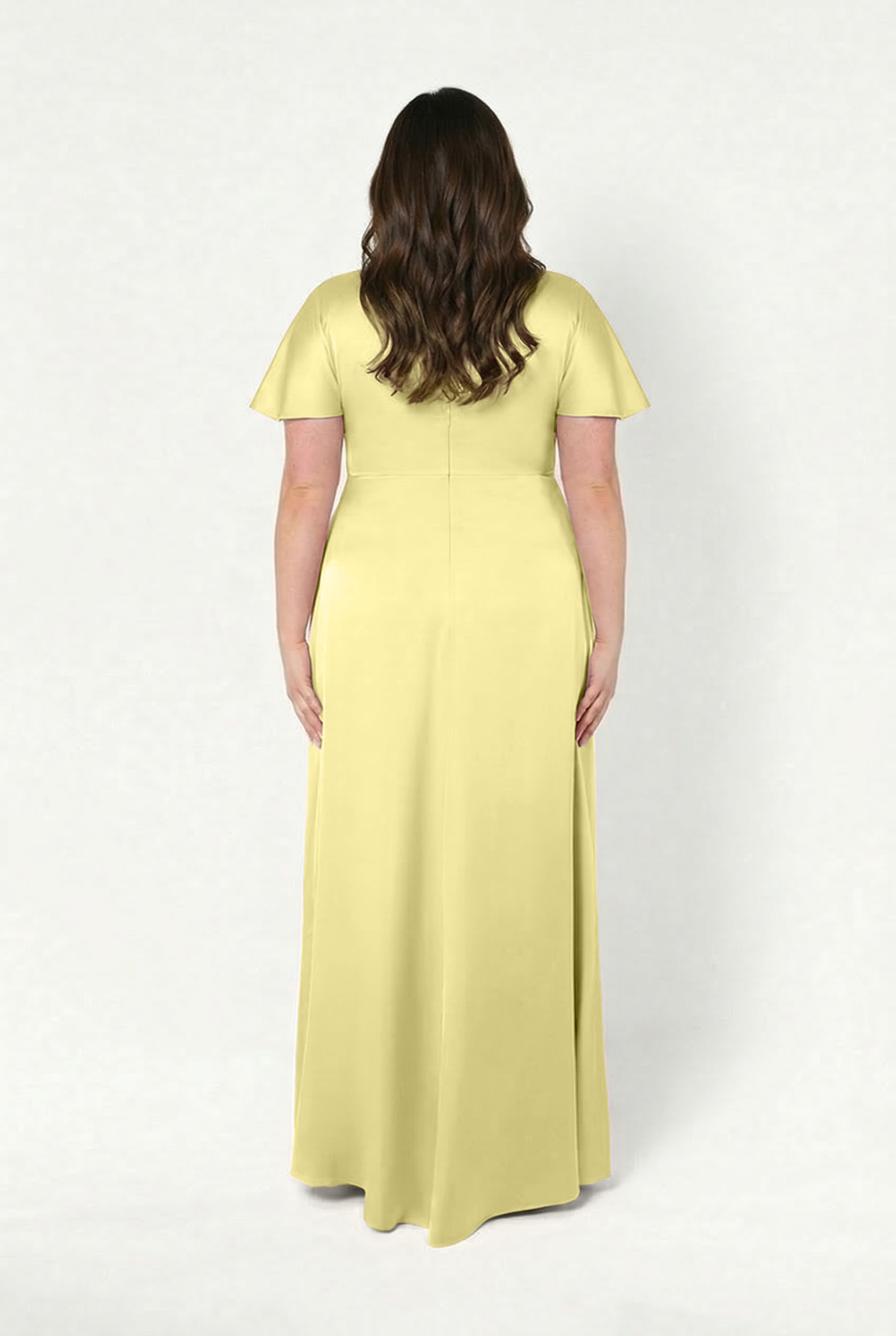 A woman with loose brown waves stands with her back to the camera, wearing the TH&TH Camilla Bridesmaid Dress in Limoncello Satin—a floor-length, size-inclusive gown with short sleeves—against a plain white background.