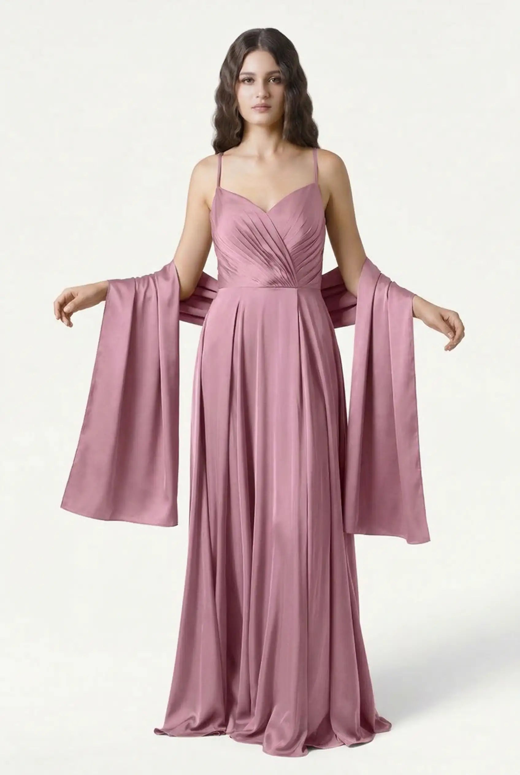 A woman in a floor-length, mauve-pink gown with thin straps and a pleated bodice stands against a plain background, elegantly holding the TH&TH Liquid Satin Shawl draped over her arms.