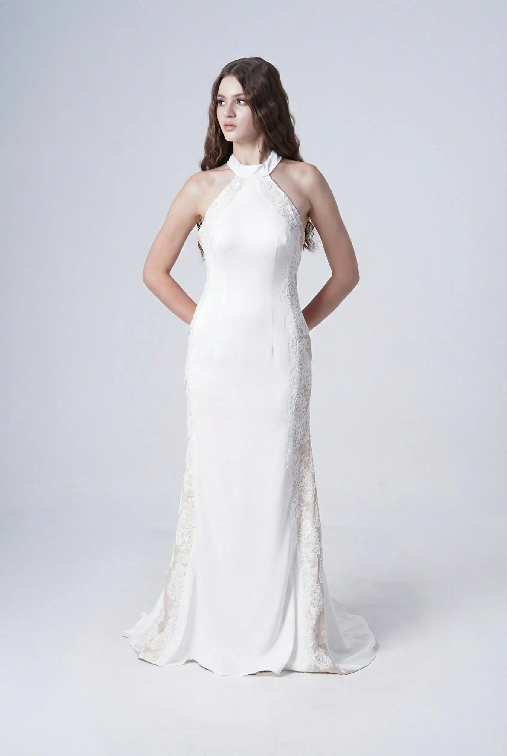 A woman with long, wavy brown hair stands against a plain background wearing the Luisa Bridal Dress in Ivory by TH&TH—a long, sleeveless satin gown with lace sides and a halter neckline, her arms behind her back.