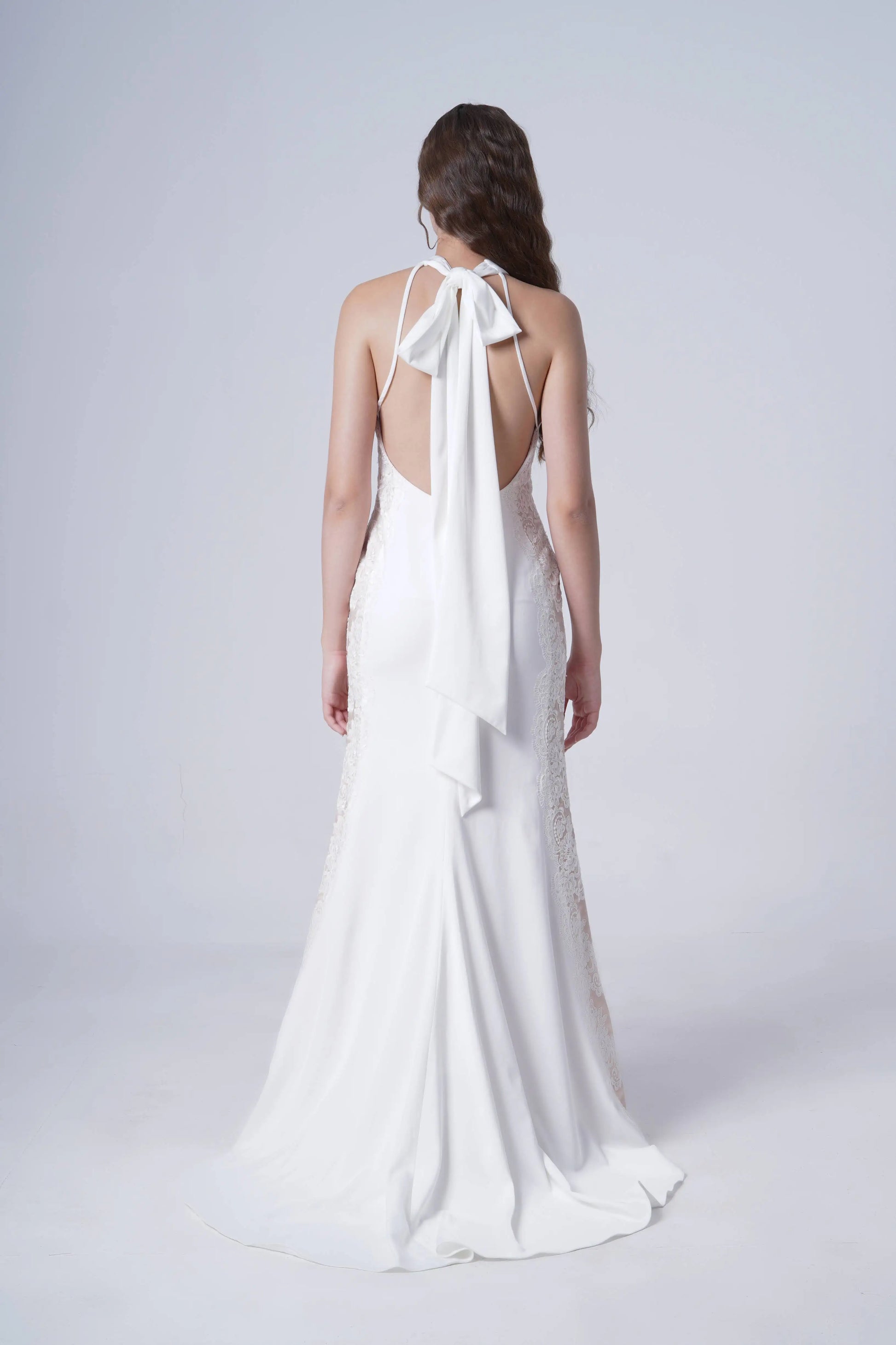 A woman with long, wavy hair faces away, wearing the Luisa Bridal Dress in Ivory by TH&TH. The satin gown features a halter neckline, a large neck bow, and a flowing train against a plain light gray background.