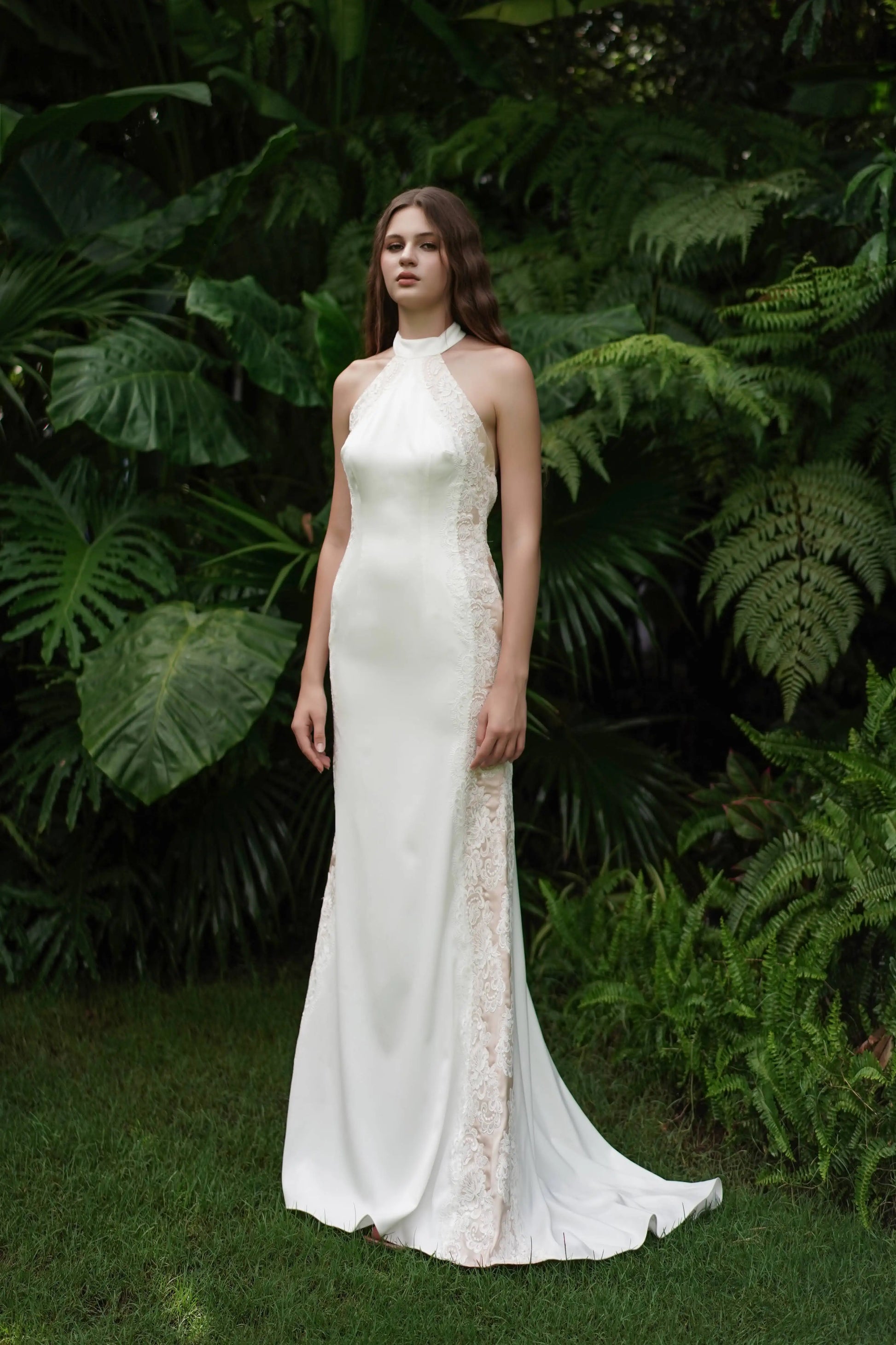 A woman stands on grass among tropical plants, wearing the Luisa Bridal Dress in Ivory by TH&TH—a sleeveless, floor-length gown with lace side panels and a halter neckline.