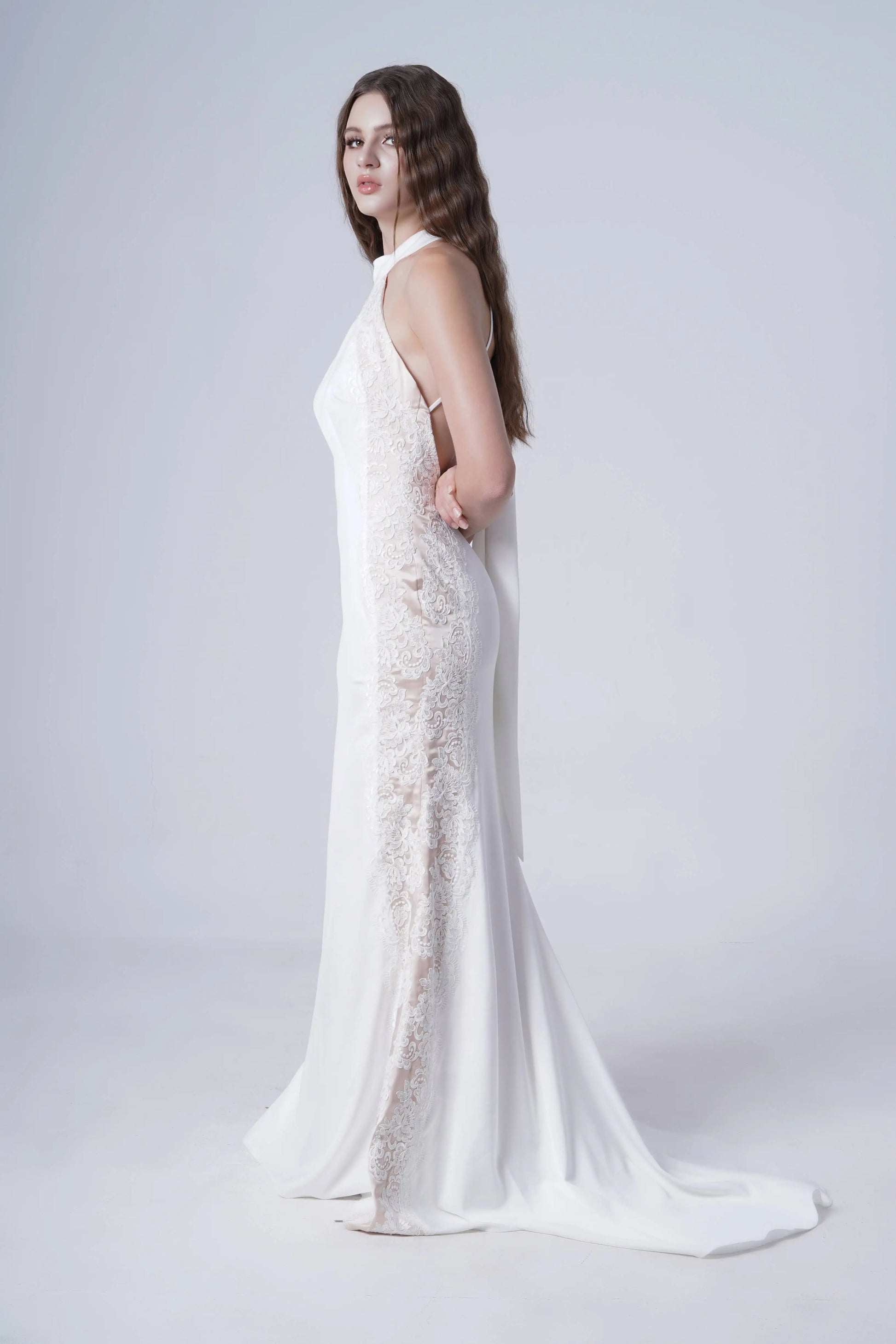 A woman with long wavy hair stands barefoot in the Luisa Bridal Dress in Ivory by TH&TH, featuring lace side panels, posing against a plain light background.