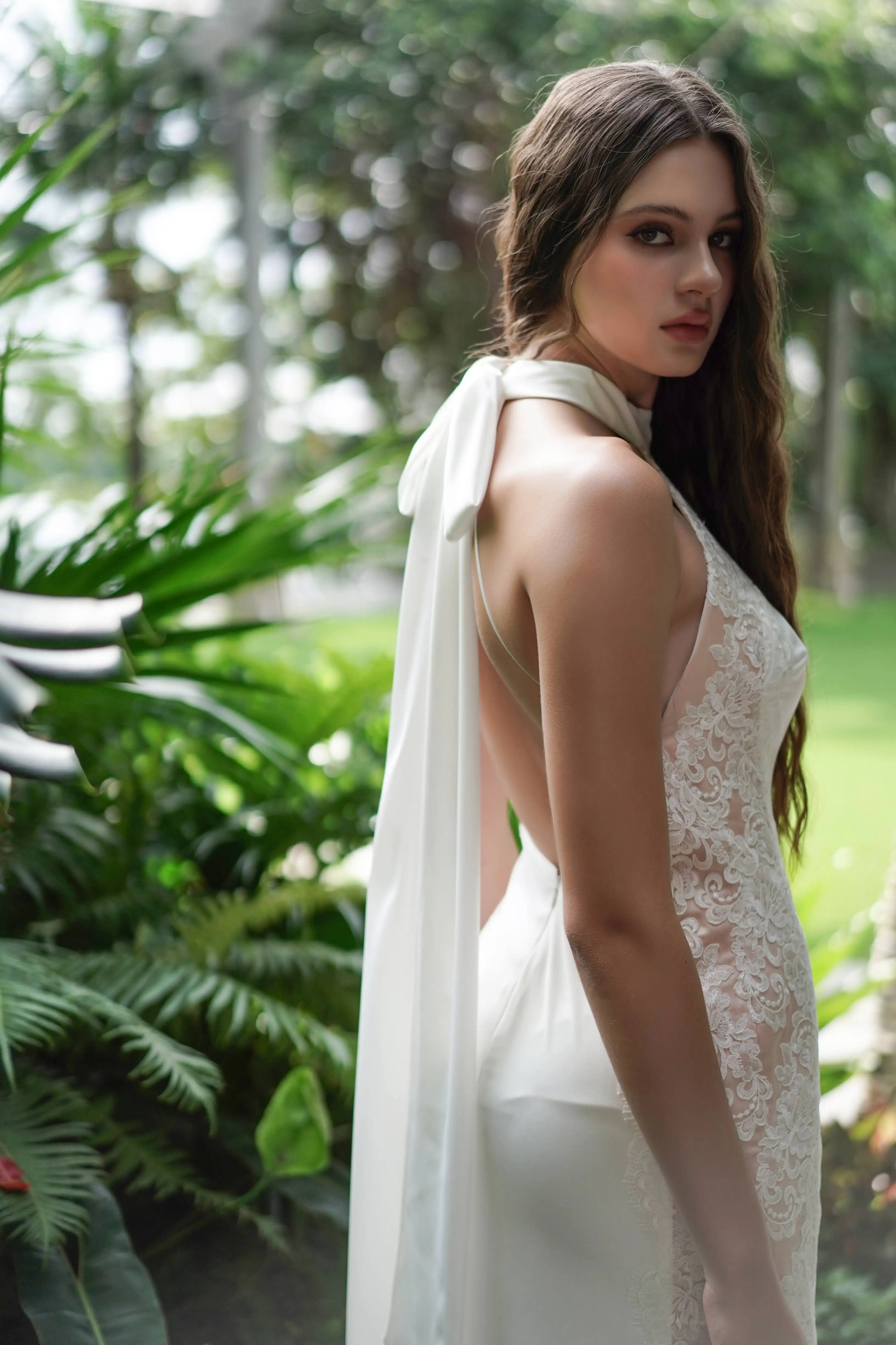 A woman in the Luisa Bridal Dress in Ivory by TH&TH, featuring a halter neckline, lace accents, and an open back with ribbon detail, stands in a lush garden, looking over her shoulder at the camera.